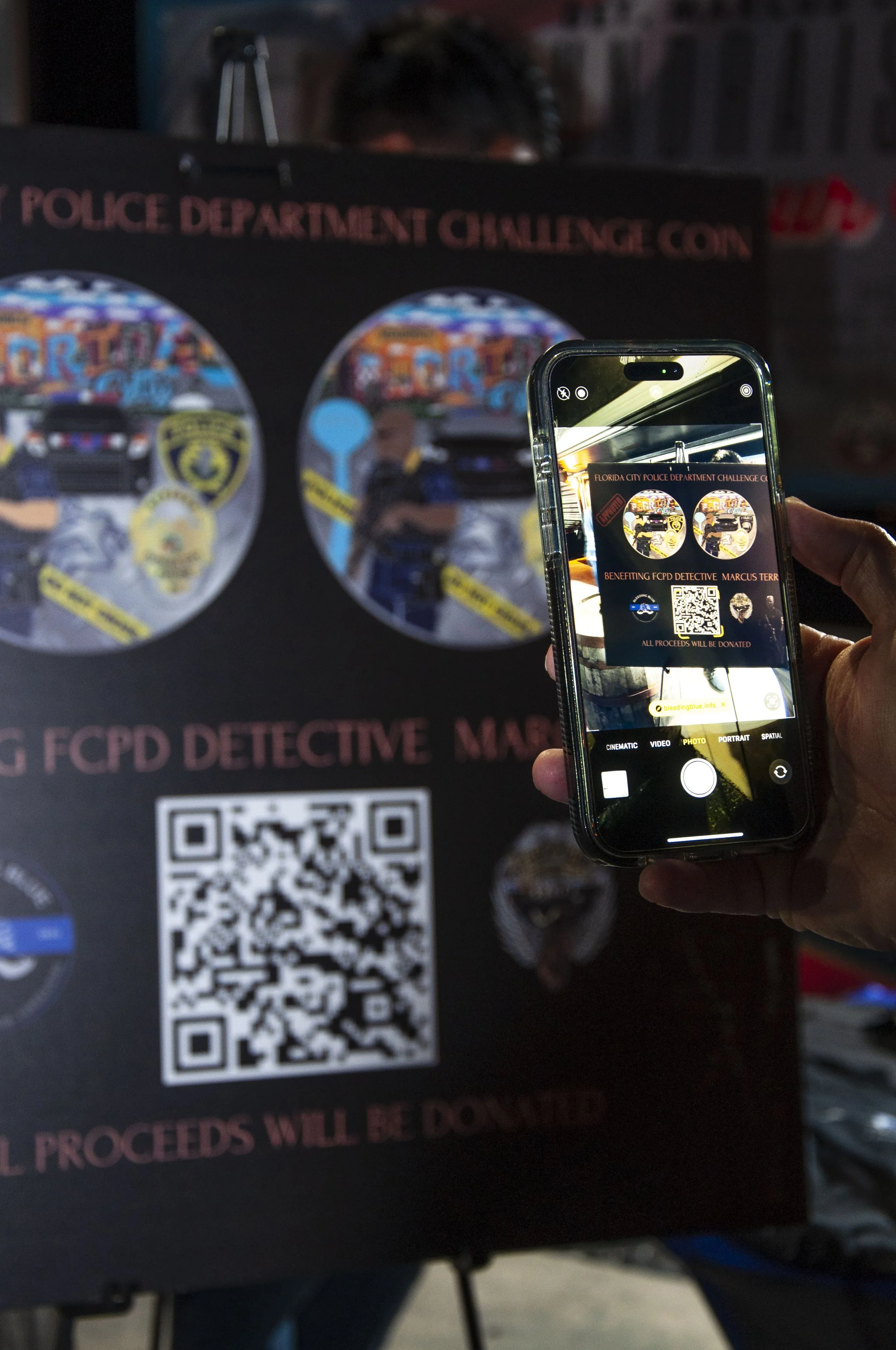 A person is holding a smartphone, taking a photo of a poster featuring police department challenge coins. The poster includes a QR code and text about fundraising for a detective. The smartphone screen shows a digital image of the poster.