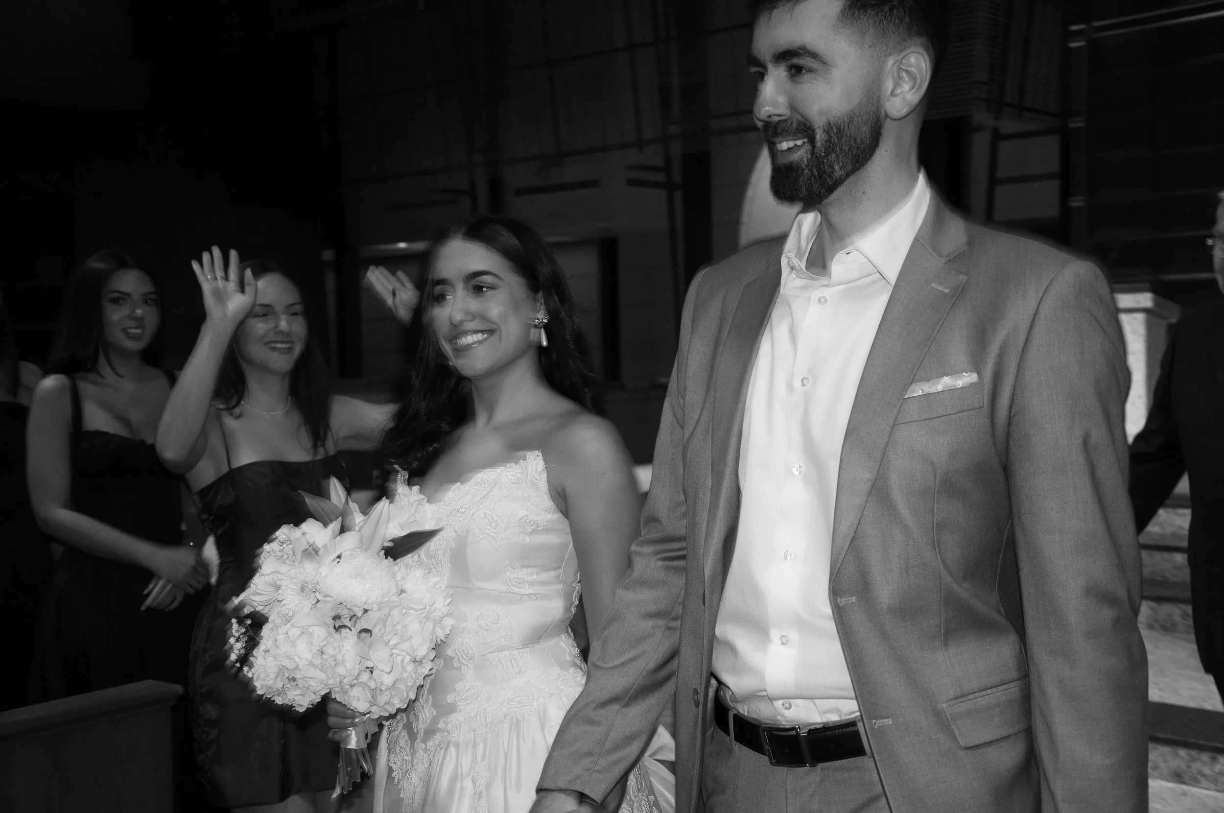 Bridal couple smiling, walking hand-in-hand; bride holding a bouquet, surrounded by bridesmaids.