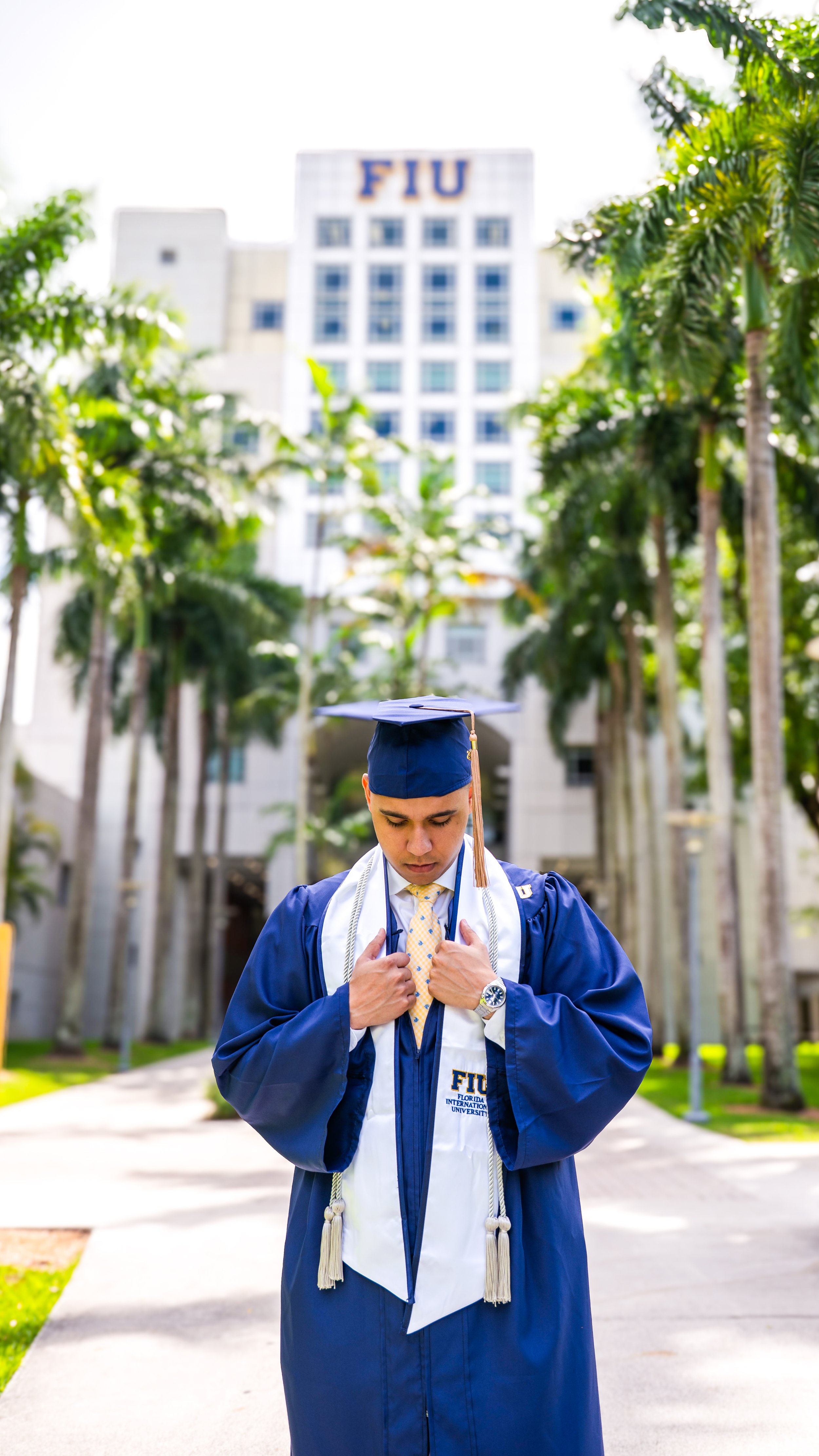 Person in blue scrubs smiling, standing outside in front of a building with "FIU" signage, palm trees, and sunlight.