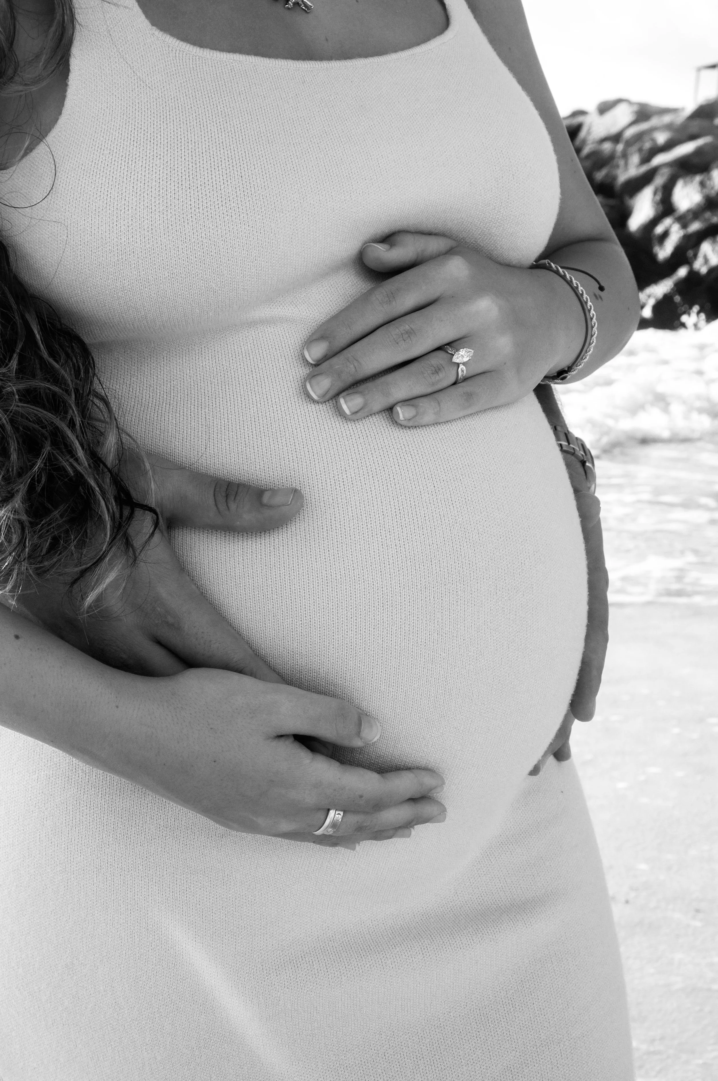 Close-up of a couple's hands on a pregnant woman's belly at the beach, with the woman wearing a tank top and rings.