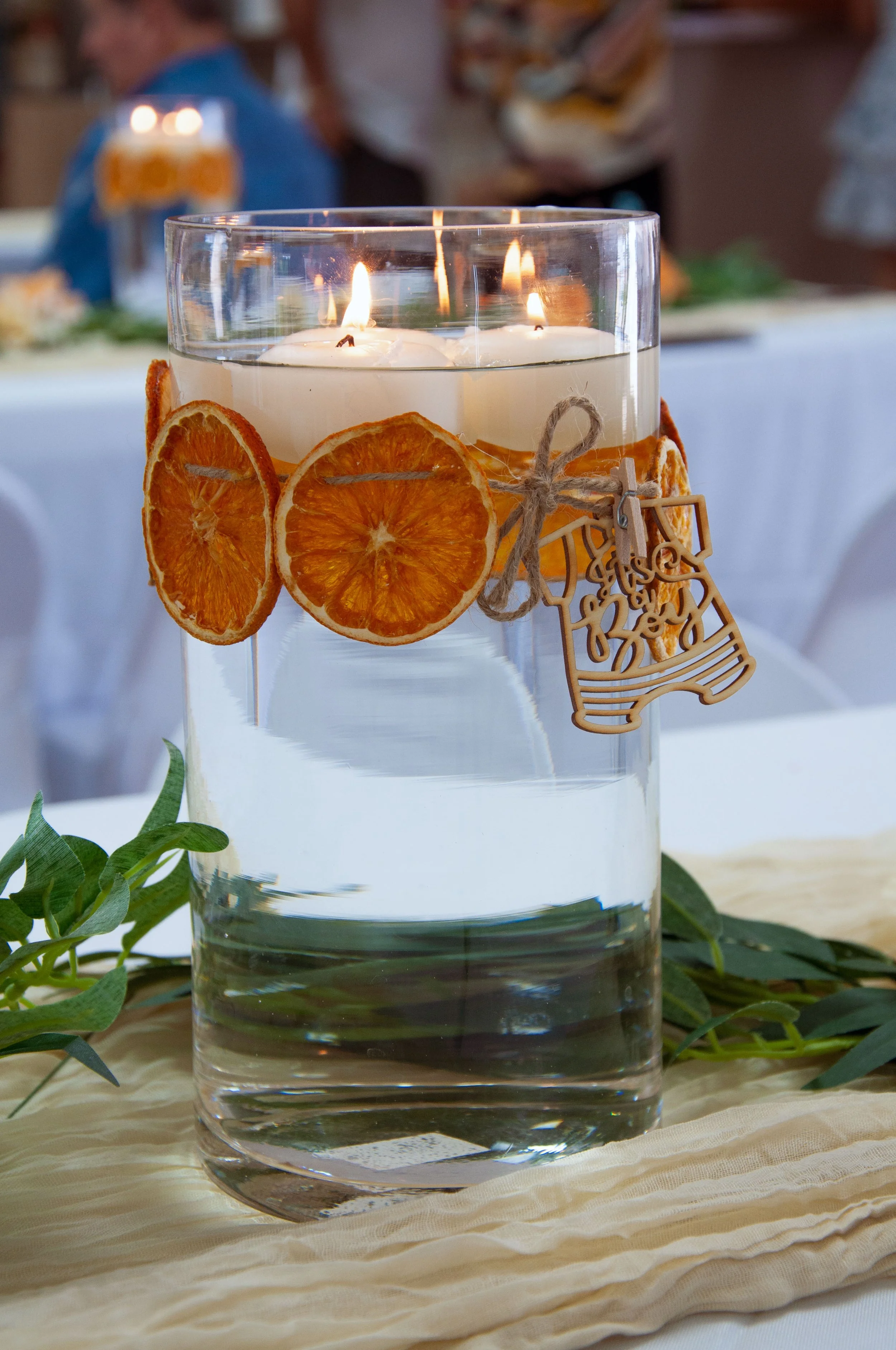 Glass vase filled with water, floating candles on top, decorated with dried orange slices, twine, and a wooden ornament, placed on a table with greenery.