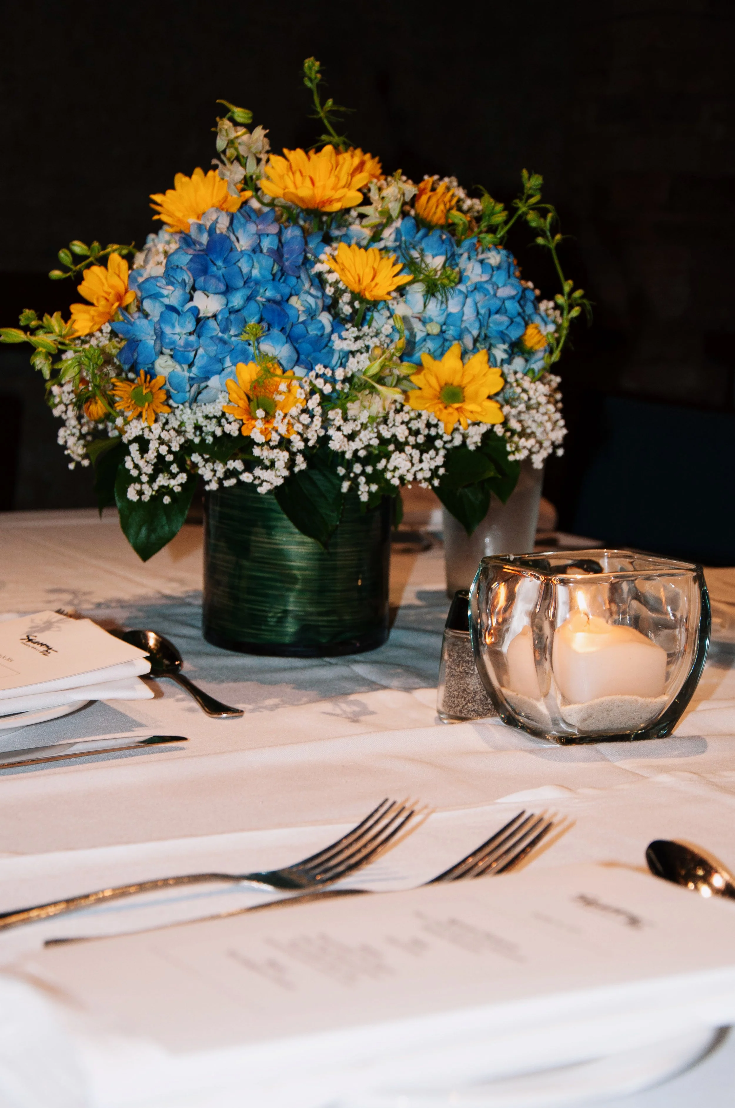 Table setting with a floral centerpiece featuring blue hydrangeas and yellow flowers, surrounded by baby's breath. A small candle in a glass holder and dining utensils are on the table.