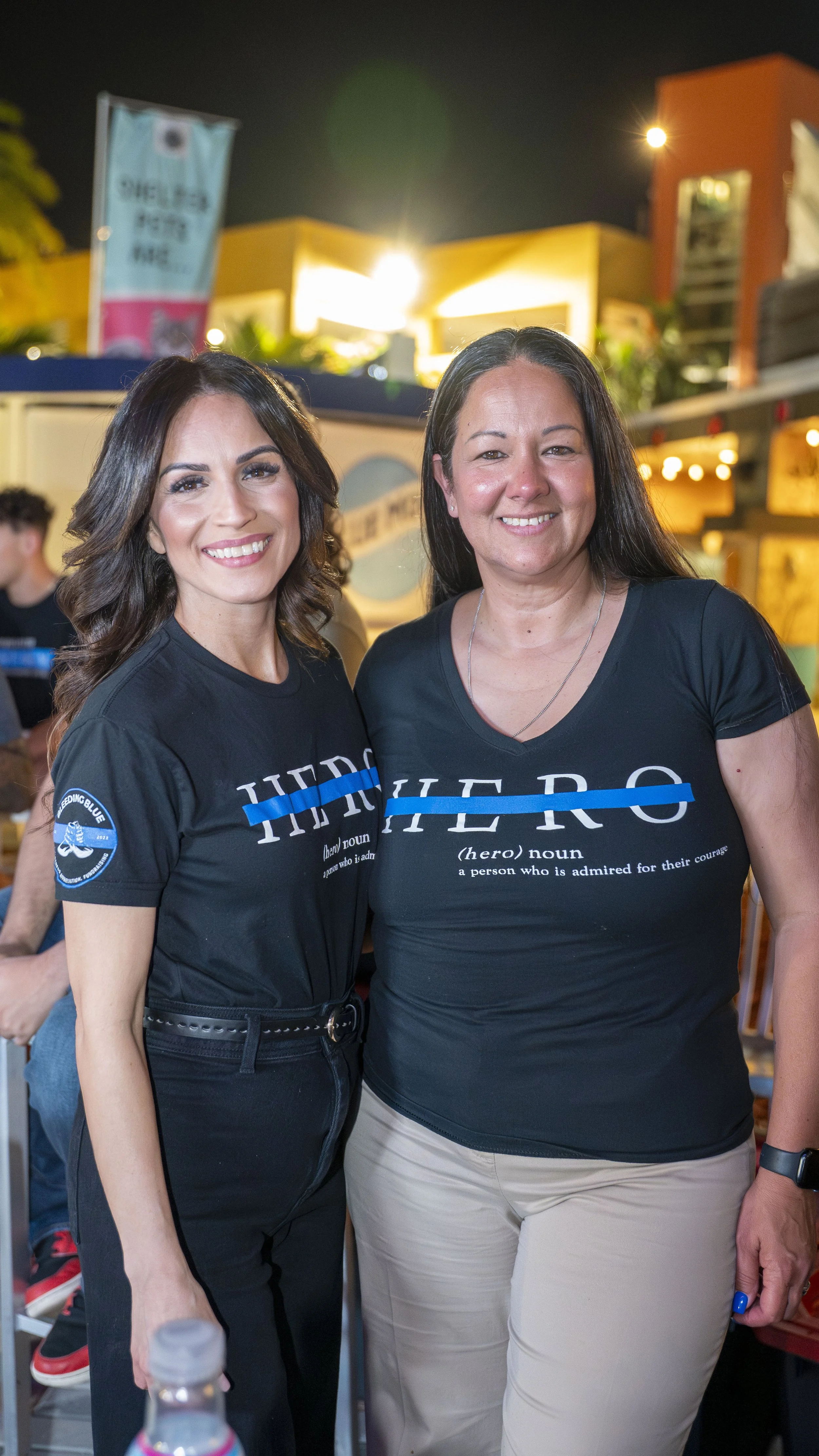 Two women smiling, wearing T-shirts with "HERO" printed on them. The background shows a nighttime outdoor setting with lights and blurred buildings.