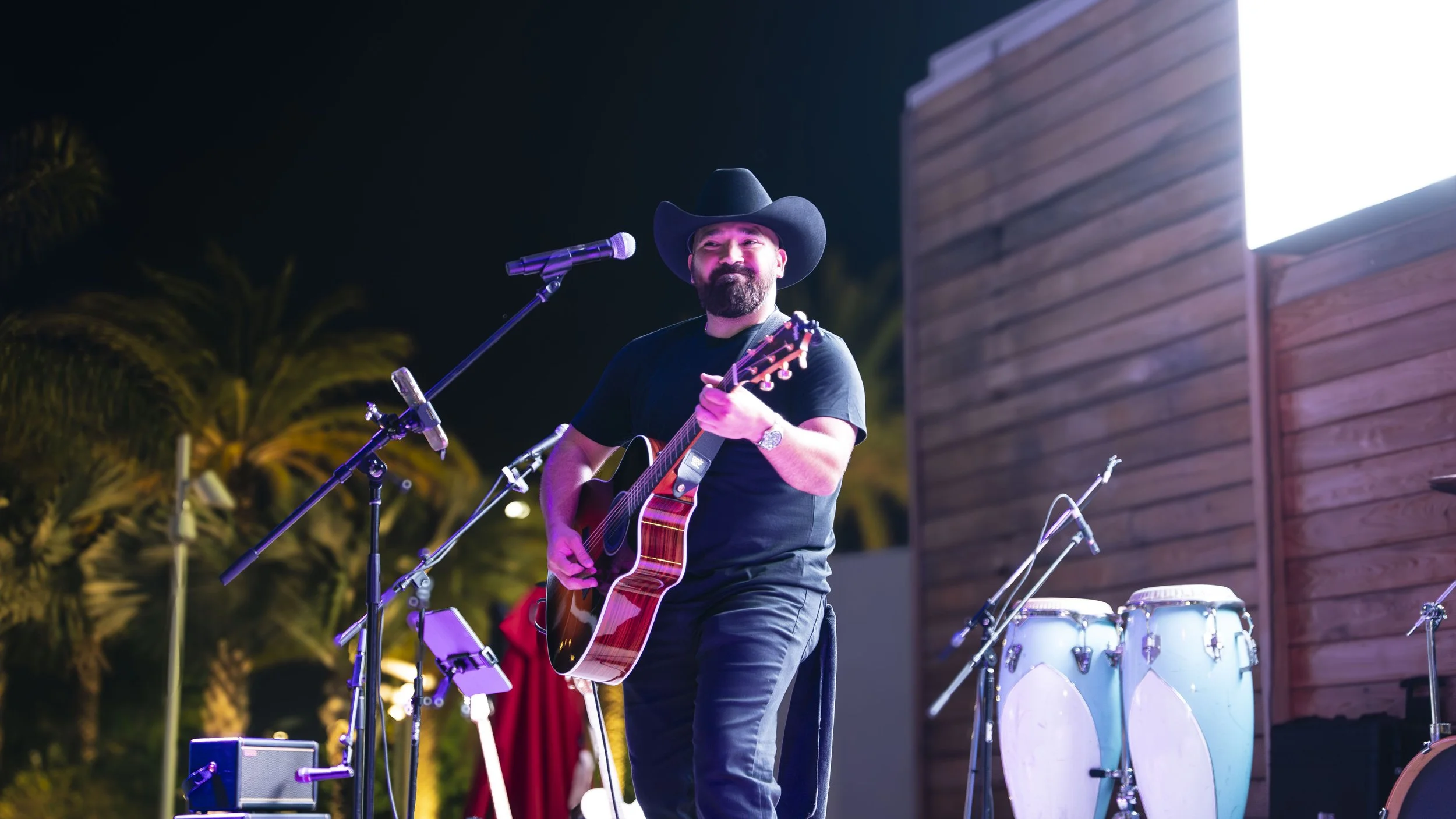 Musician playing acoustic guitar on stage at night wearing a cowboy hat, with conga drums and palm trees in the background.