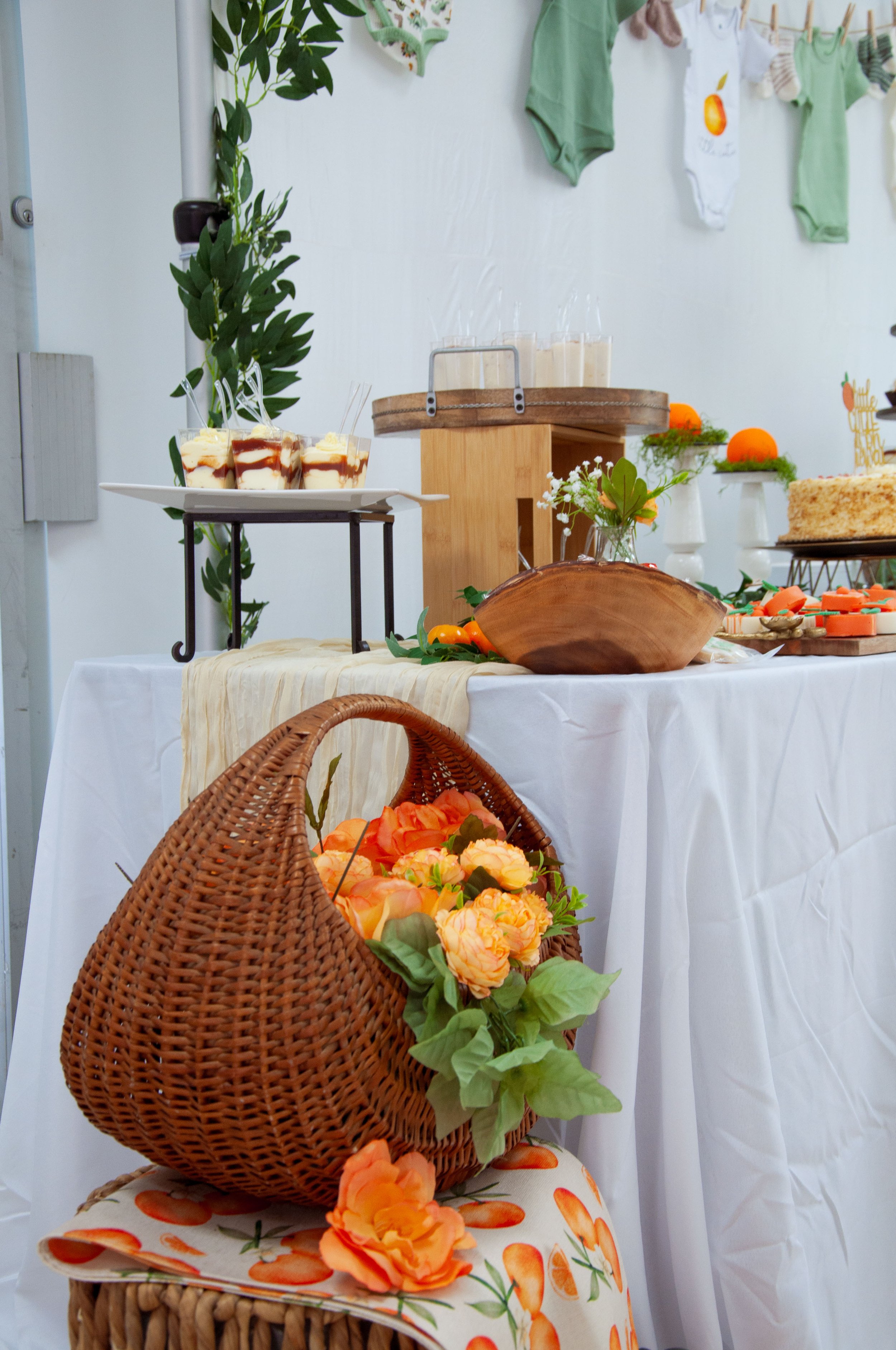 Baby shower dessert table with wicker basket, orange flowers, and baby clothes.