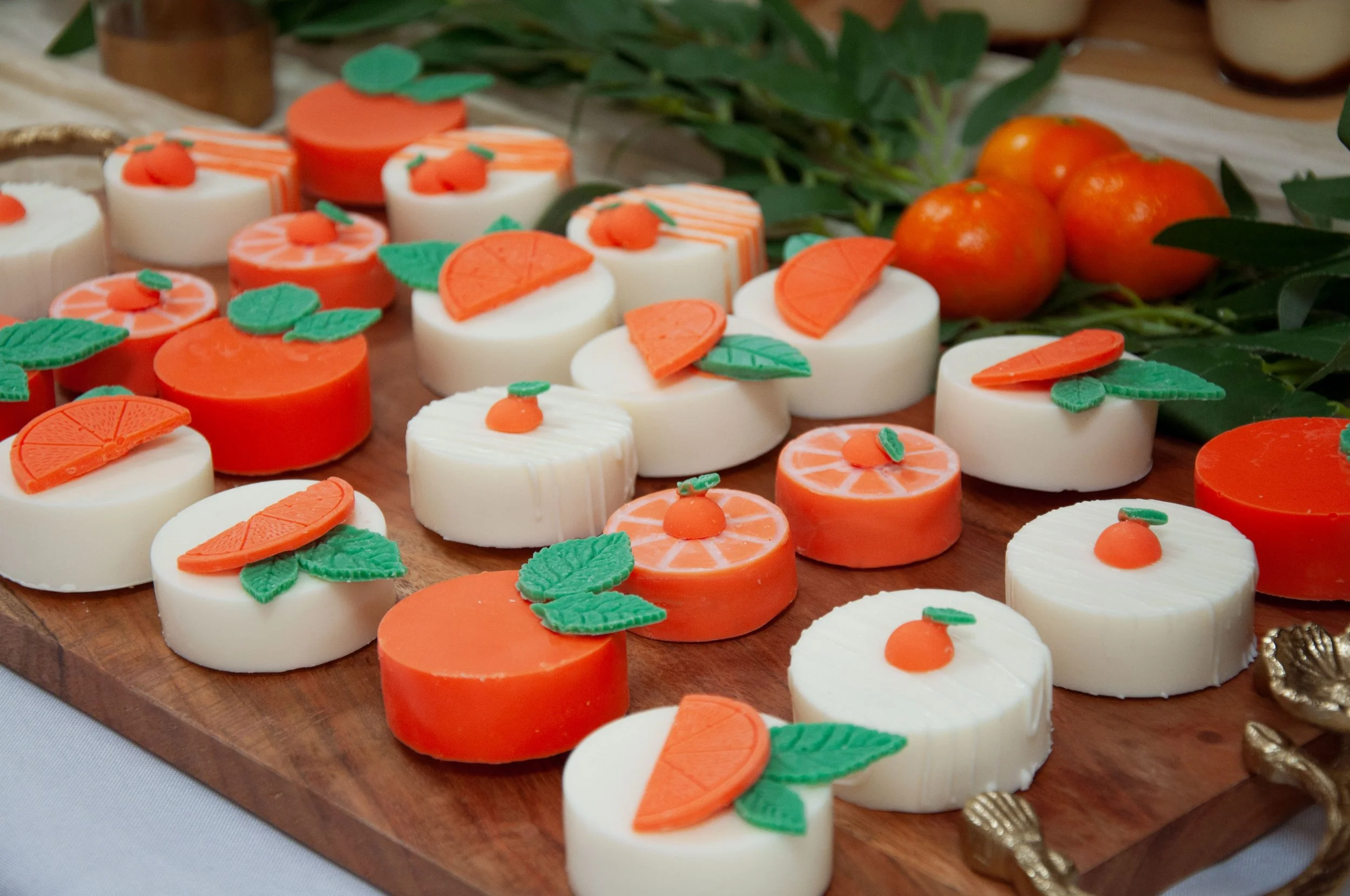 Assorted orange and white circular desserts, decorated with fondant oranges and leaves, arranged on a wooden board with tangerines and greenery in the background.