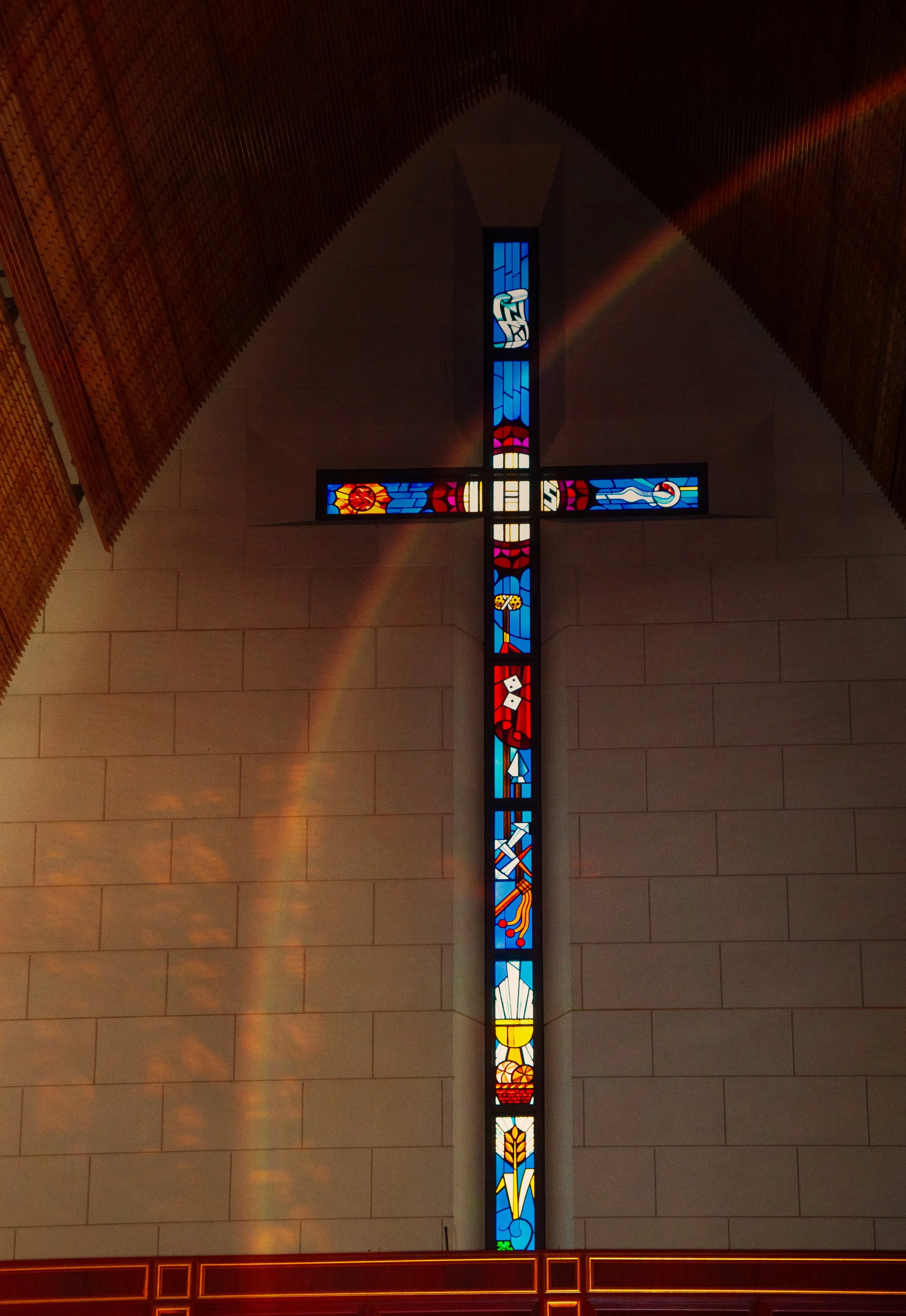 Stained glass cross in a church wall, depicting religious symbols with a rainbow effect.