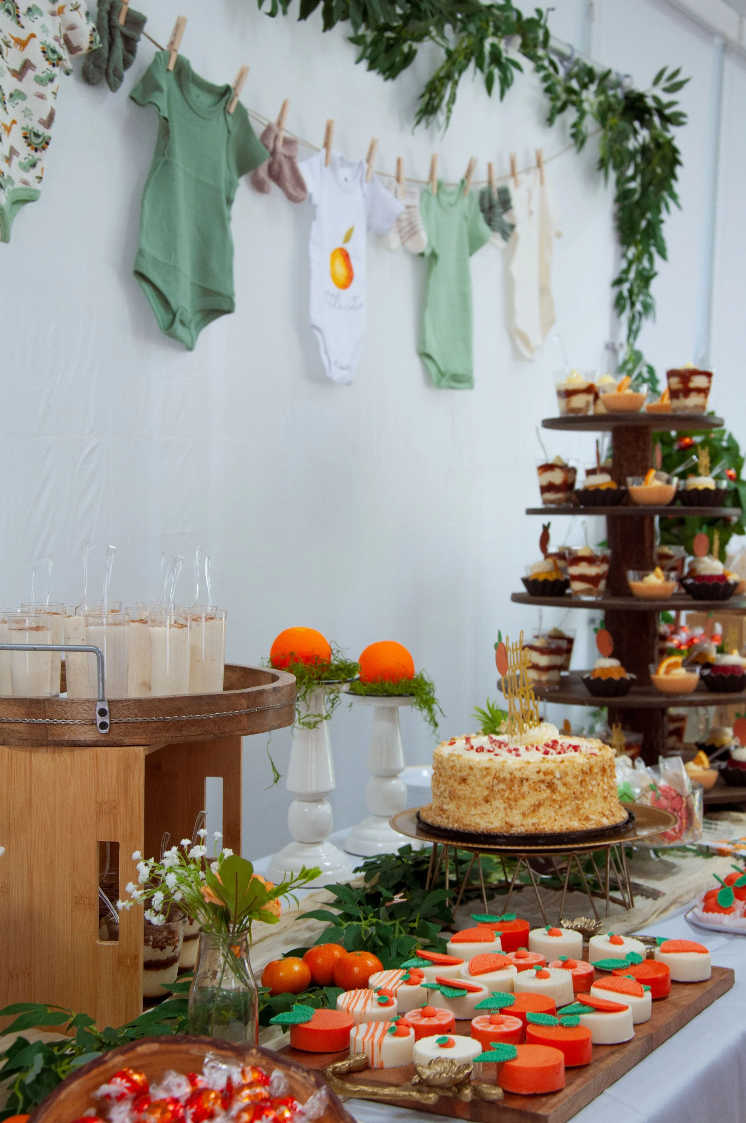 Baby shower dessert table with cake, various sweets, oranges, and decorations like baby onesies and greenery.