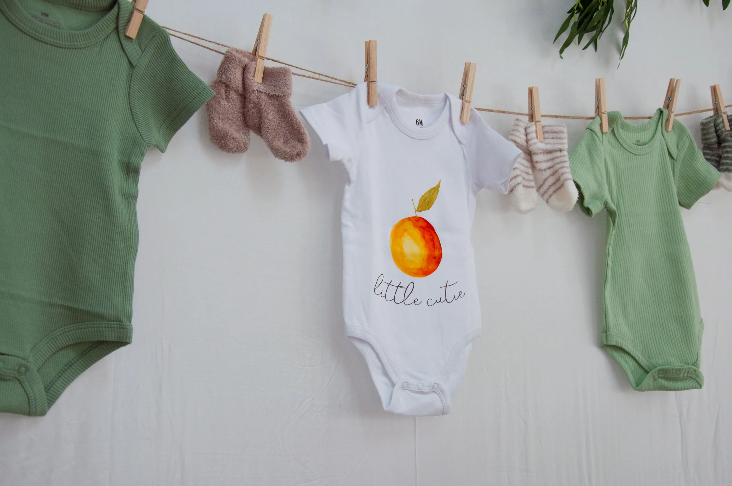 Baby clothes and socks hanging on a clothesline; includes a white onesie with a fruit graphic and the words "little cutie," green onesies, and striped socks.