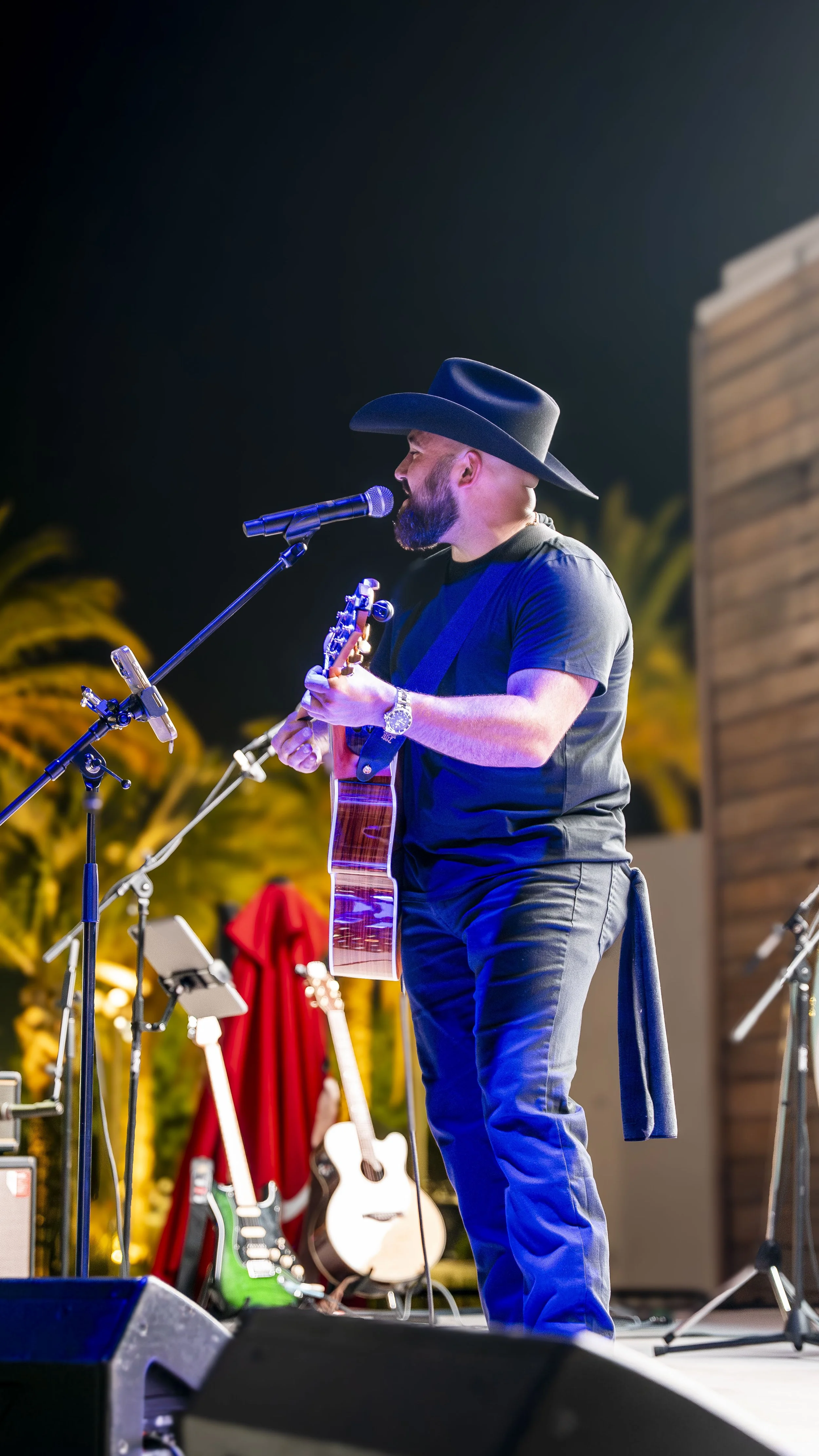 Musician wearing a black hat and shirt playing an acoustic guitar and singing into a microphone on stage.