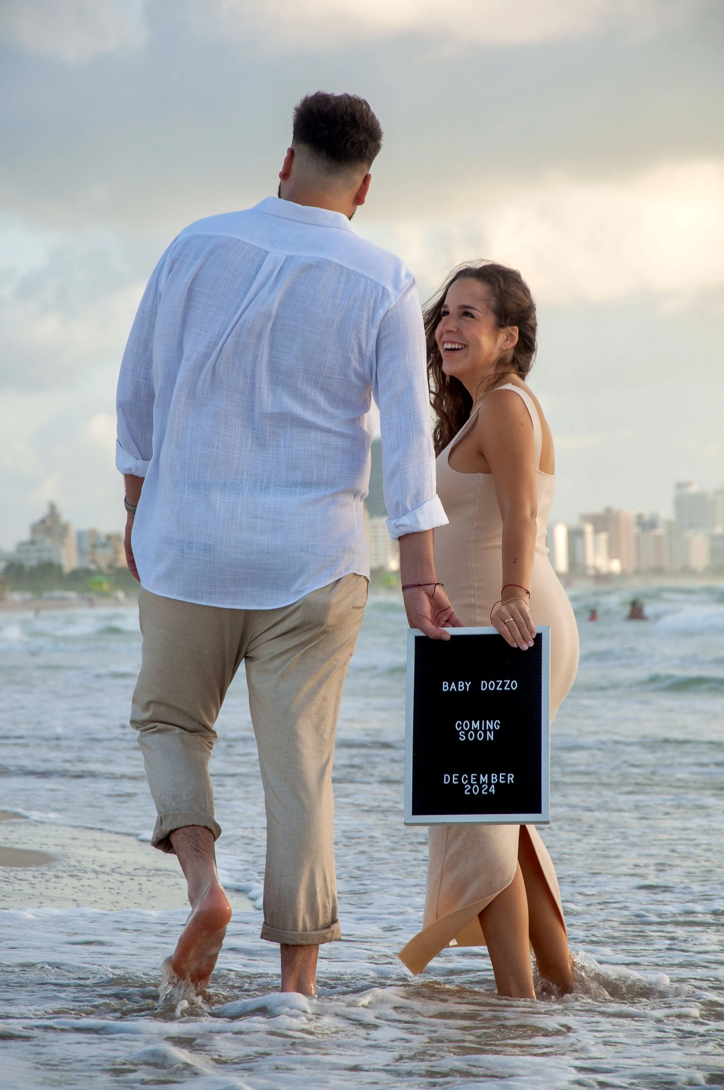 Couple walking on the beach, woman holding a sign announcing pregnancy, text reading "Baby Dozzo Coming Soon December 2024."