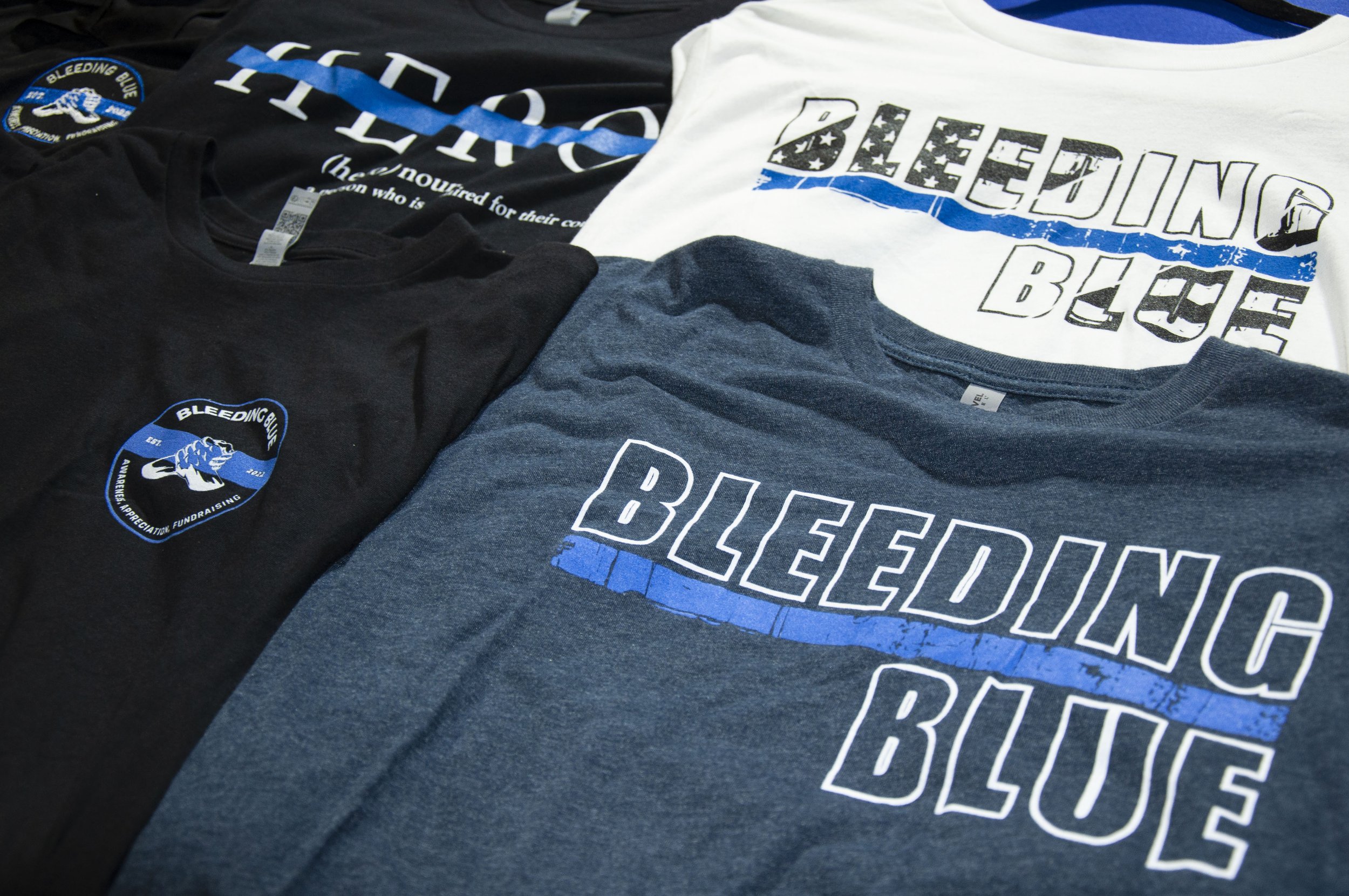Four T-shirts with "Bleeding Blue" and "Hero" designs, in black, white, and blue, featuring supportive law enforcement themes.