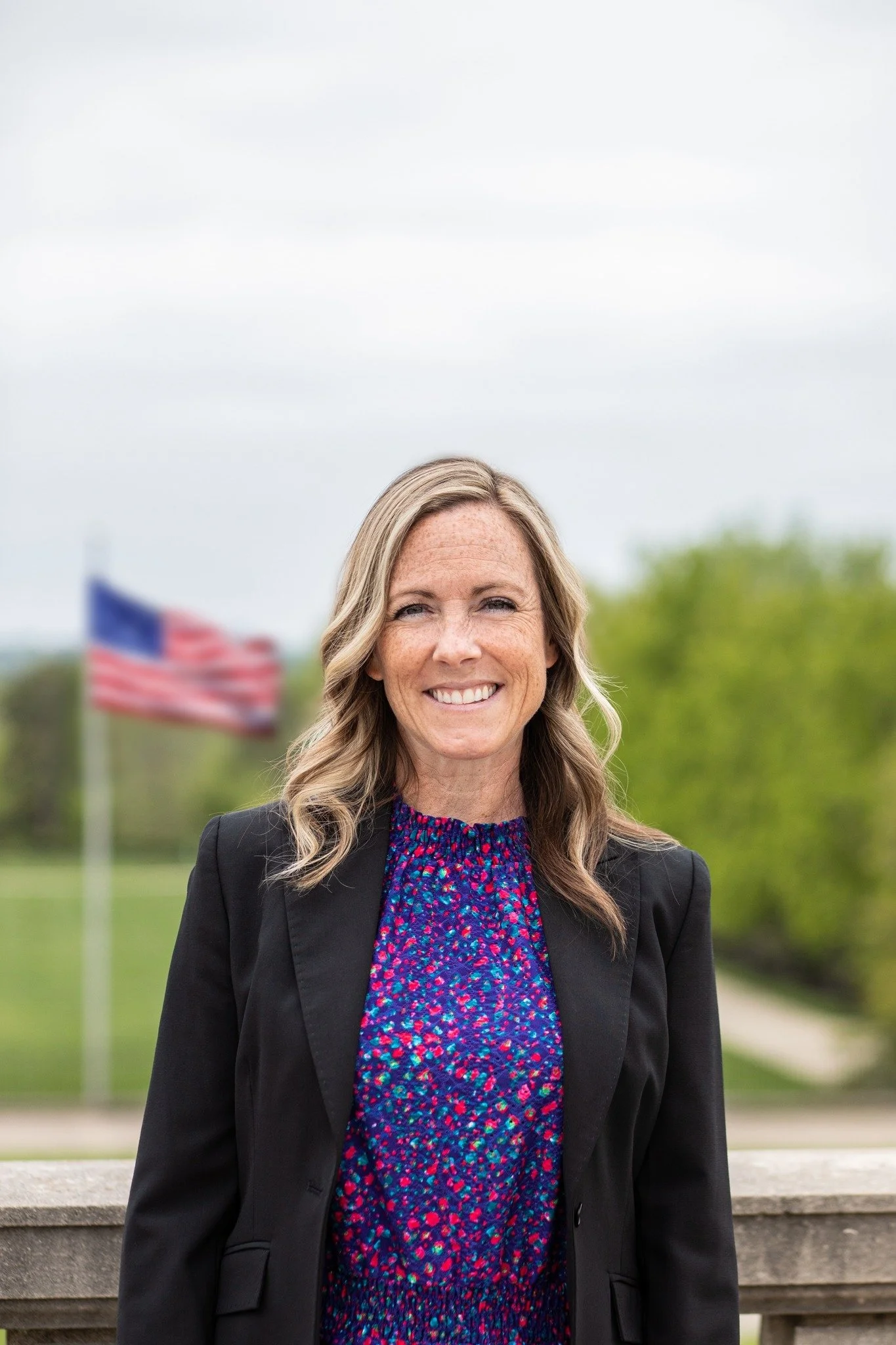 A woman with blonde hair smiling outdoors, with American flags and greenery in the background.