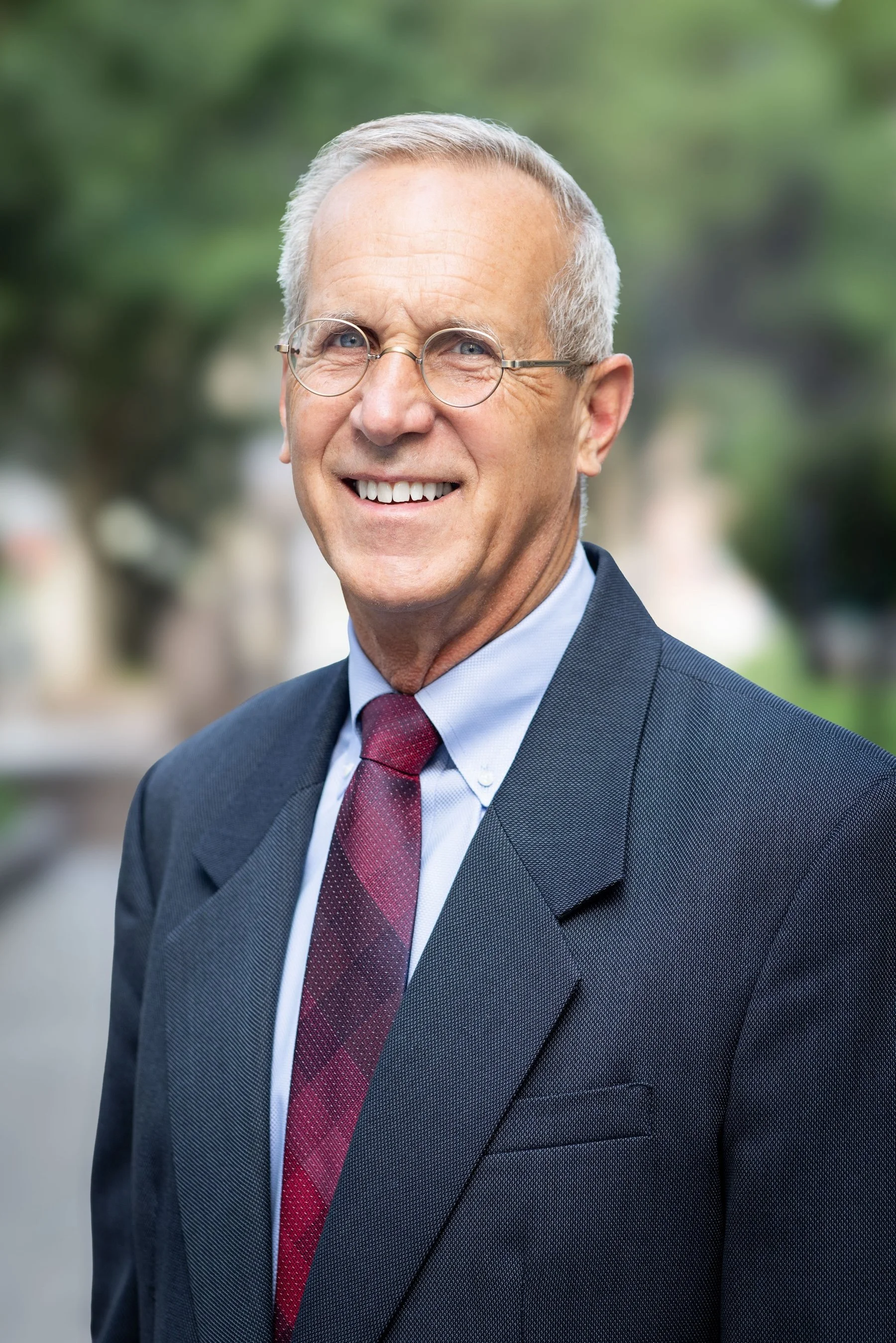 Smiling elderly man with glasses in a suit outdoors with trees in the background.