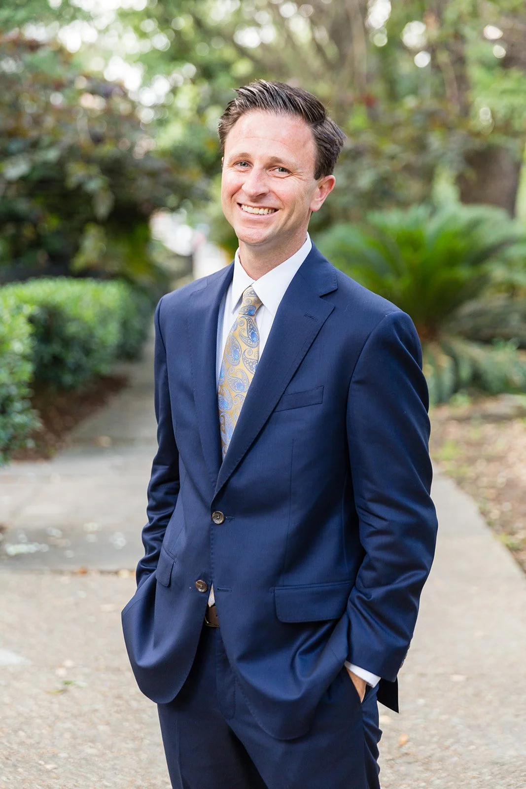 A man in a dark blue suit and a patterned tie standing outdoors on a sidewalk, smiling.