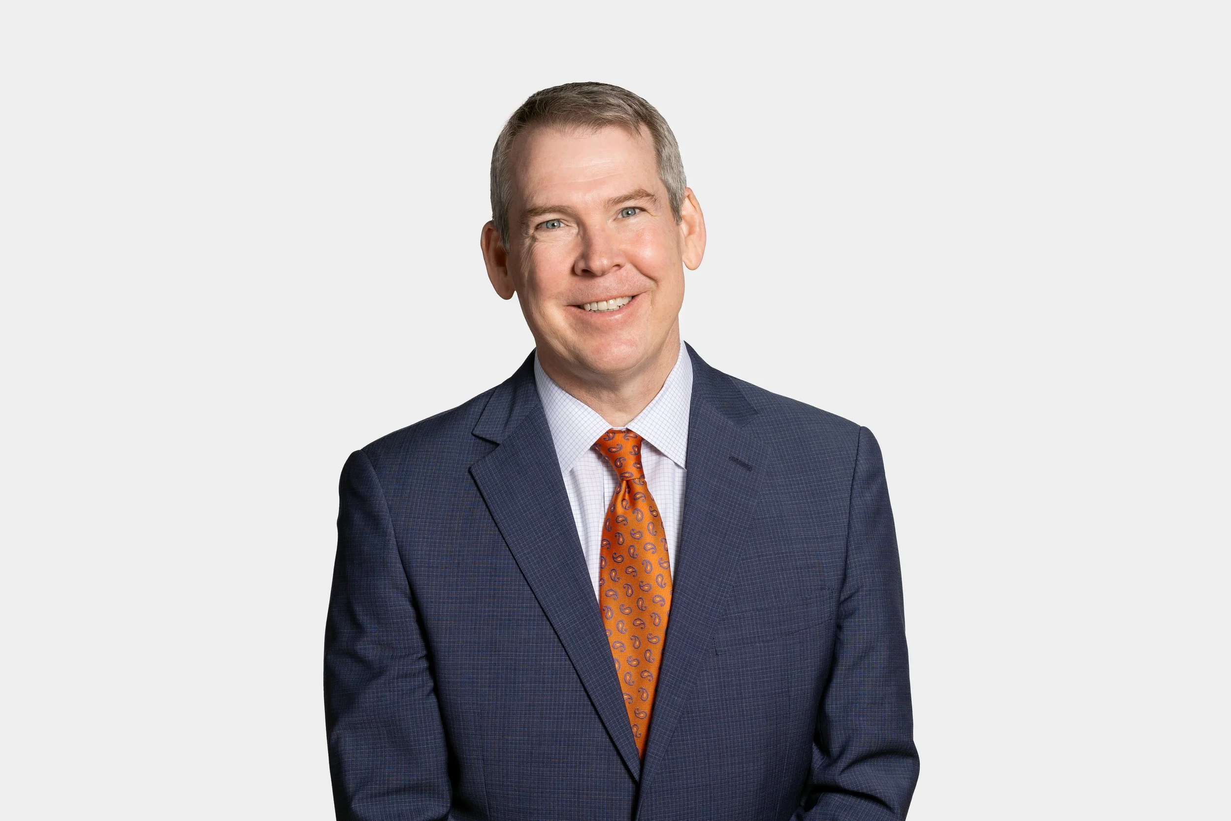 A smiling man in a dark blue plaid suit, white dress shirt, and orange paisley tie posing against a plain white background.