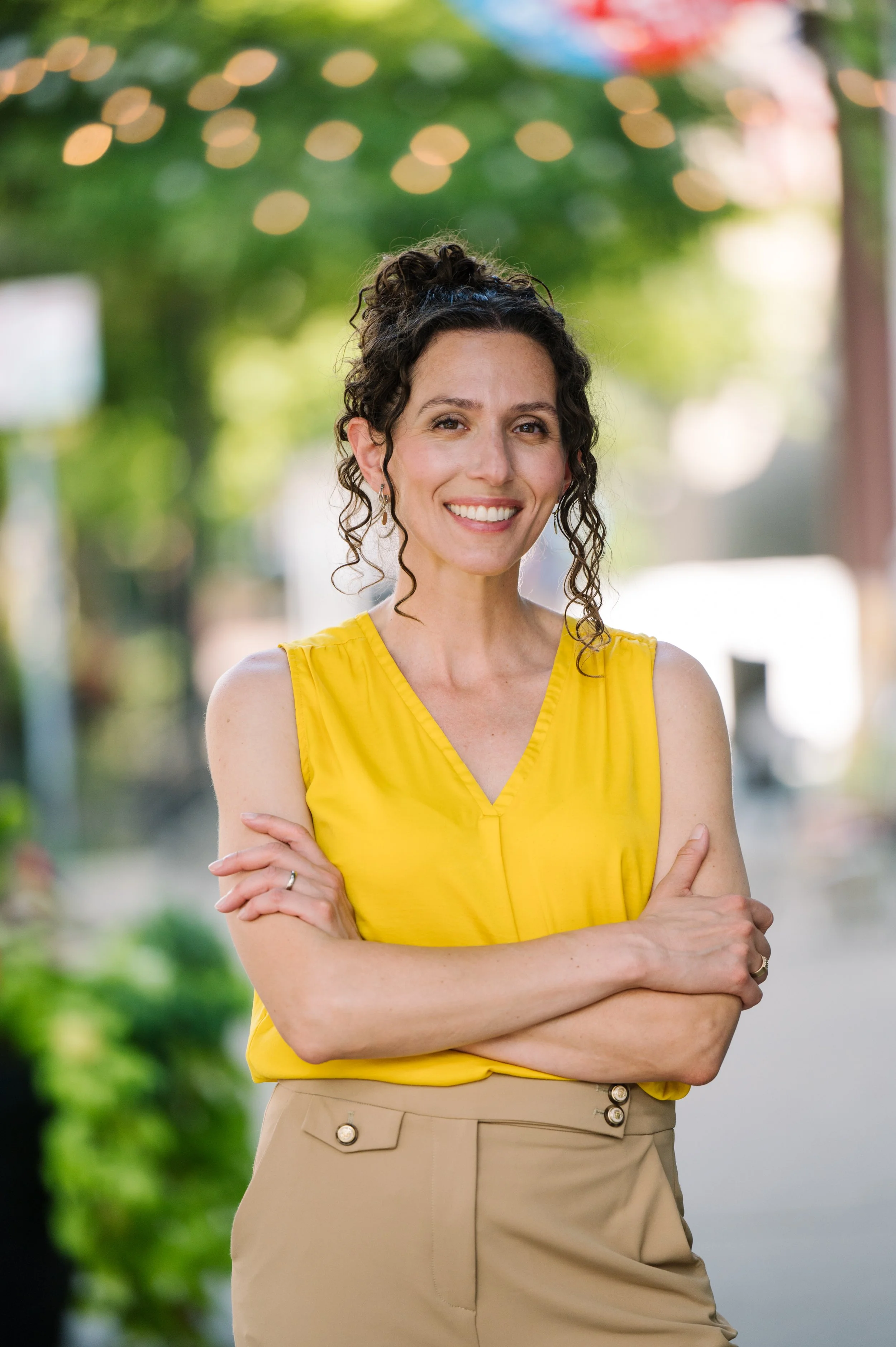 A woman with curly dark hair smiling, wearing a yellow sleeveless top and beige pants, standing outdoors on a sunny day with blurred greenery and string lights in the background.