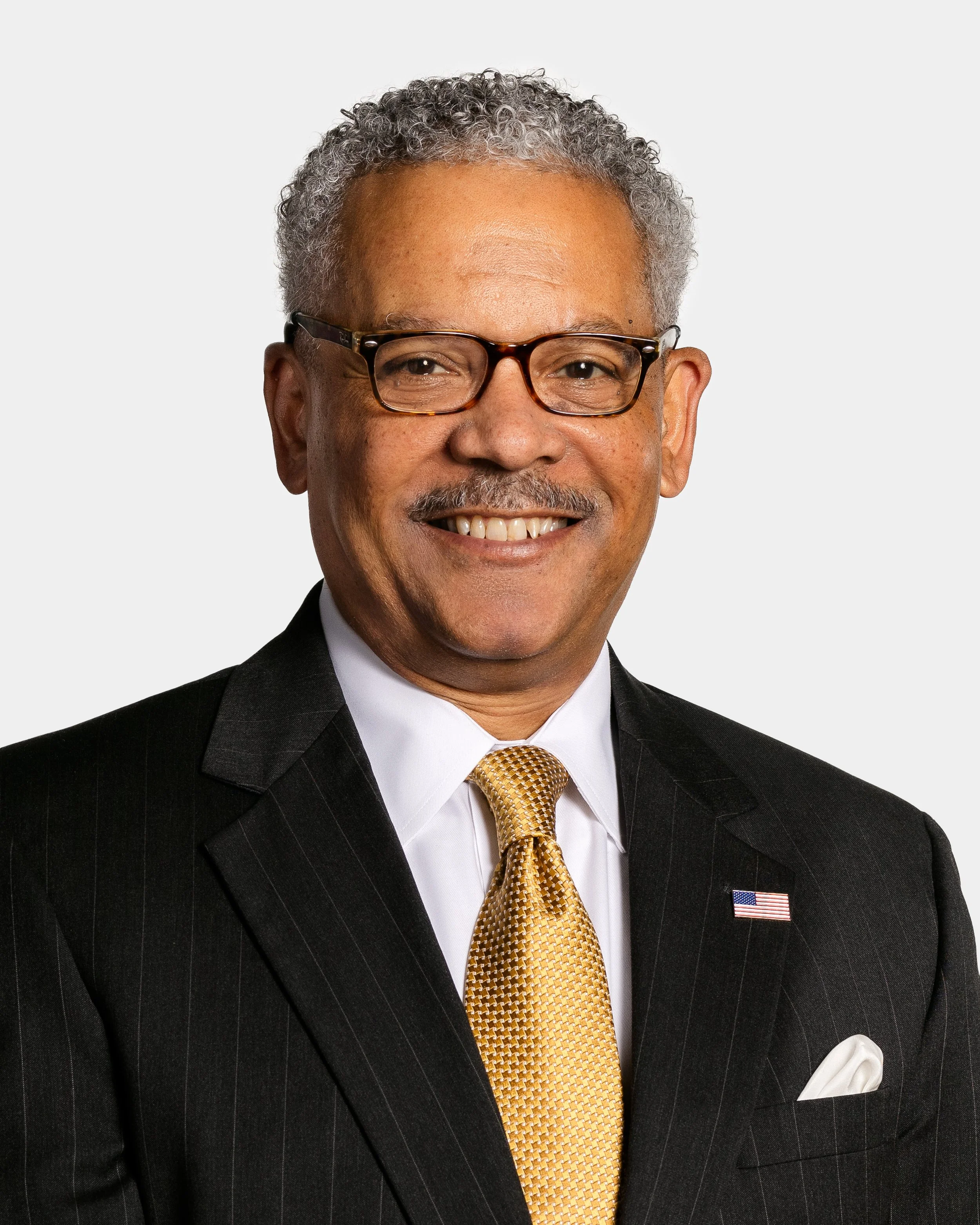 Smiling middle-aged man in black suit, white shirt, yellow tie, glasses, with an American flag pin on his lapel, against a plain white background.