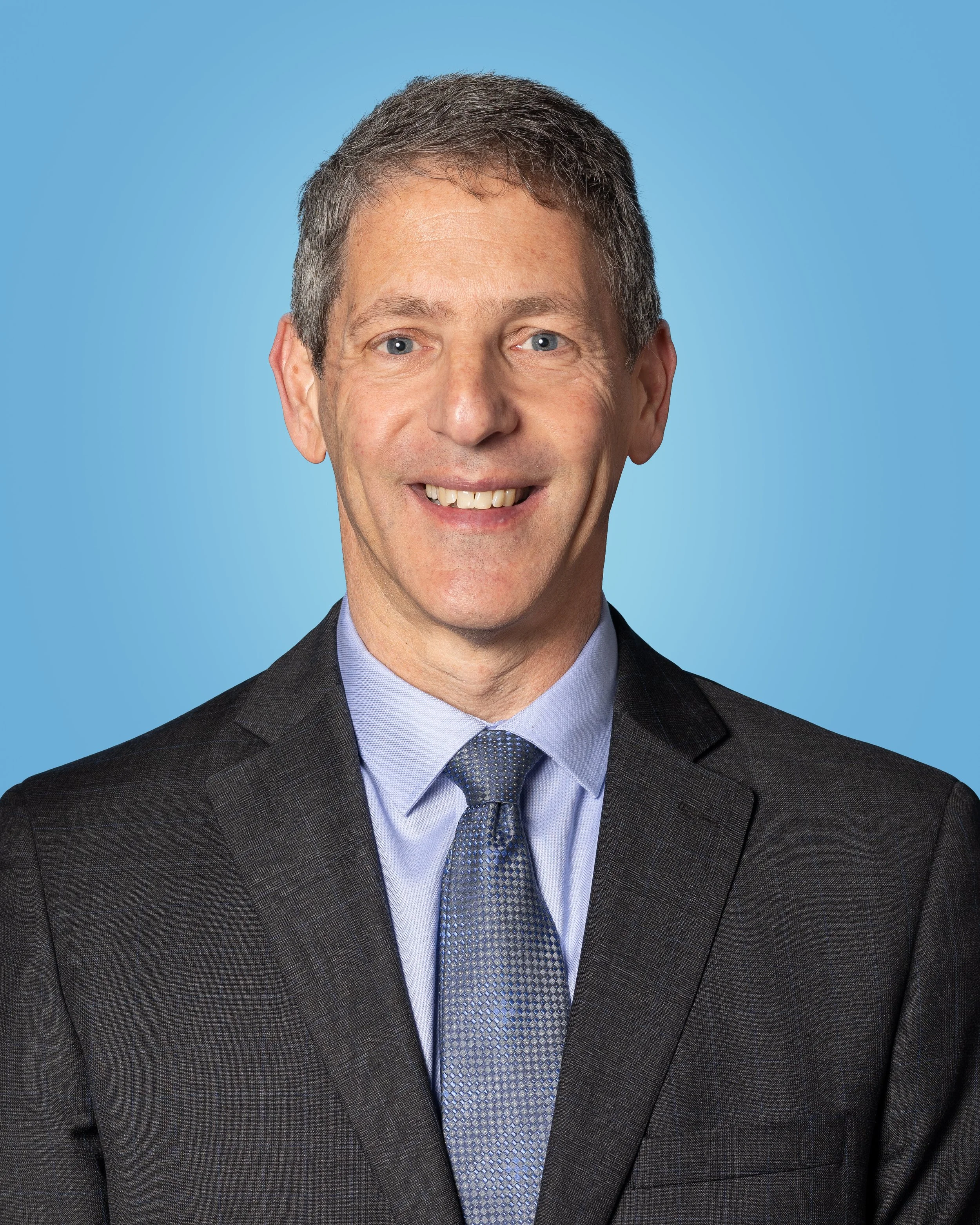A man in a dark business suit, light blue shirt, and patterned tie, smiling at the camera against a blue background.