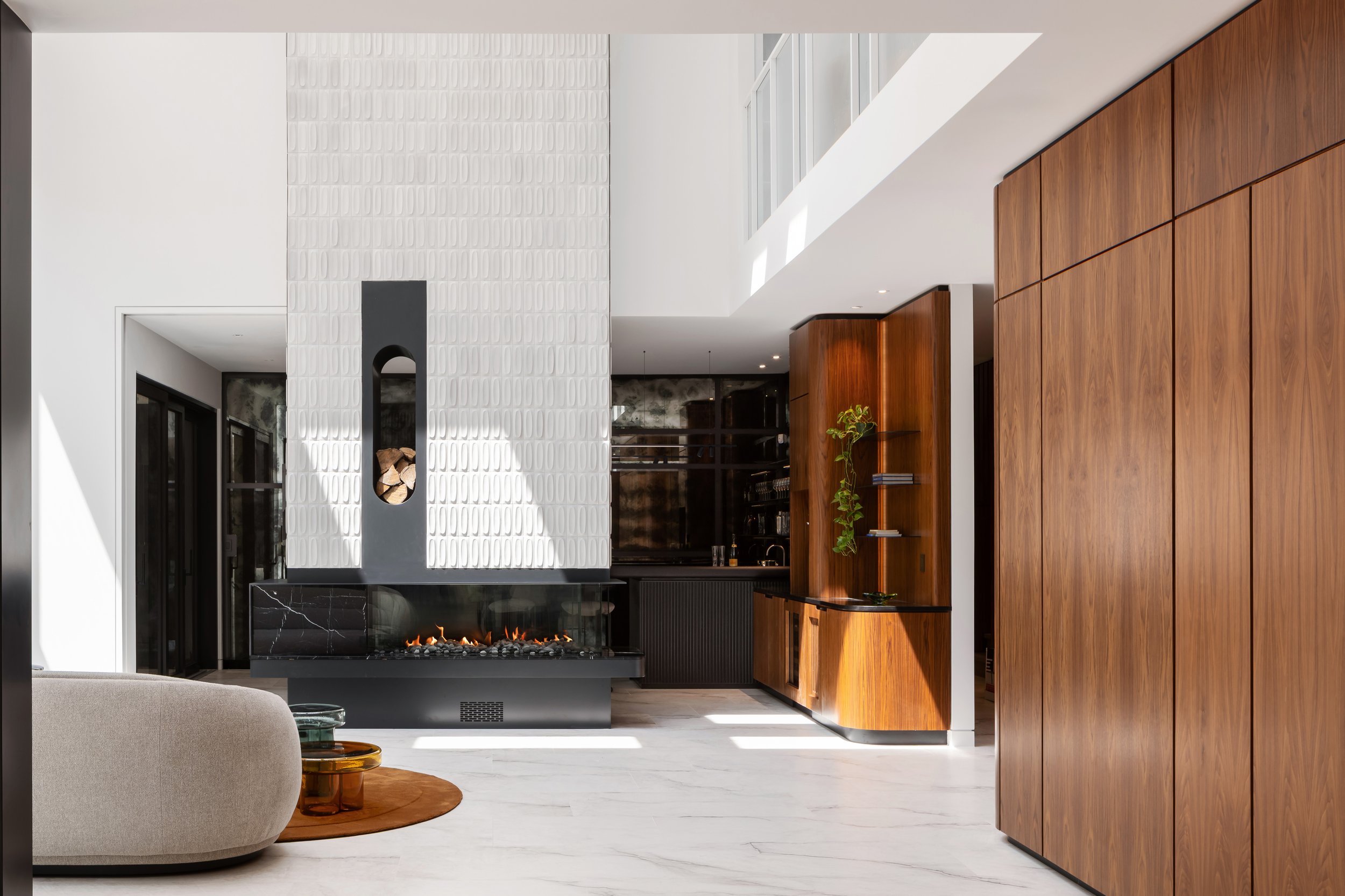 Modern living room with a fireplace, white 3D textured tiled wall, wooden cabinetry, and large windows letting in natural light.