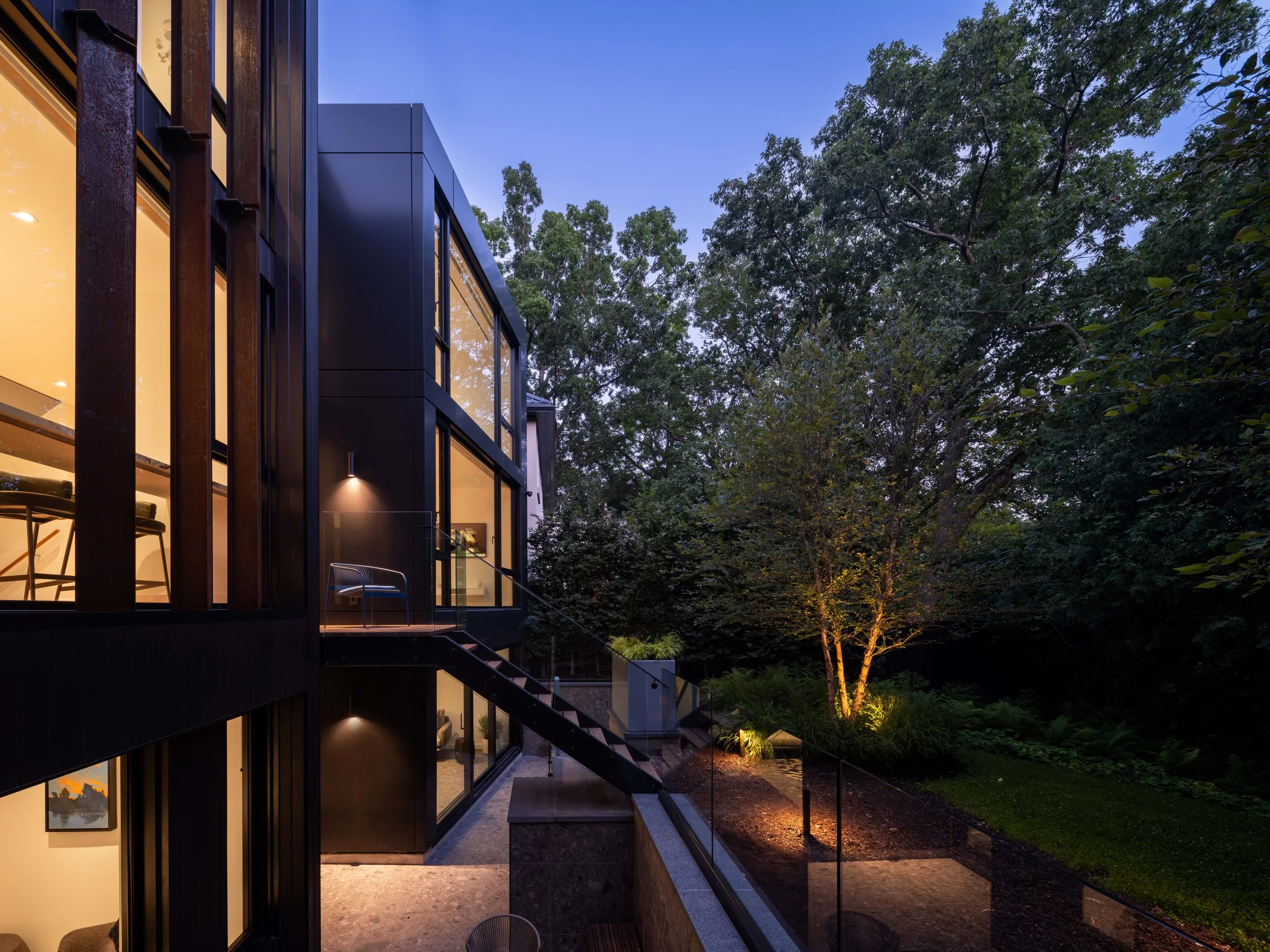 Modern house with glass windows illuminated from the inside, surrounded by trees at dusk.