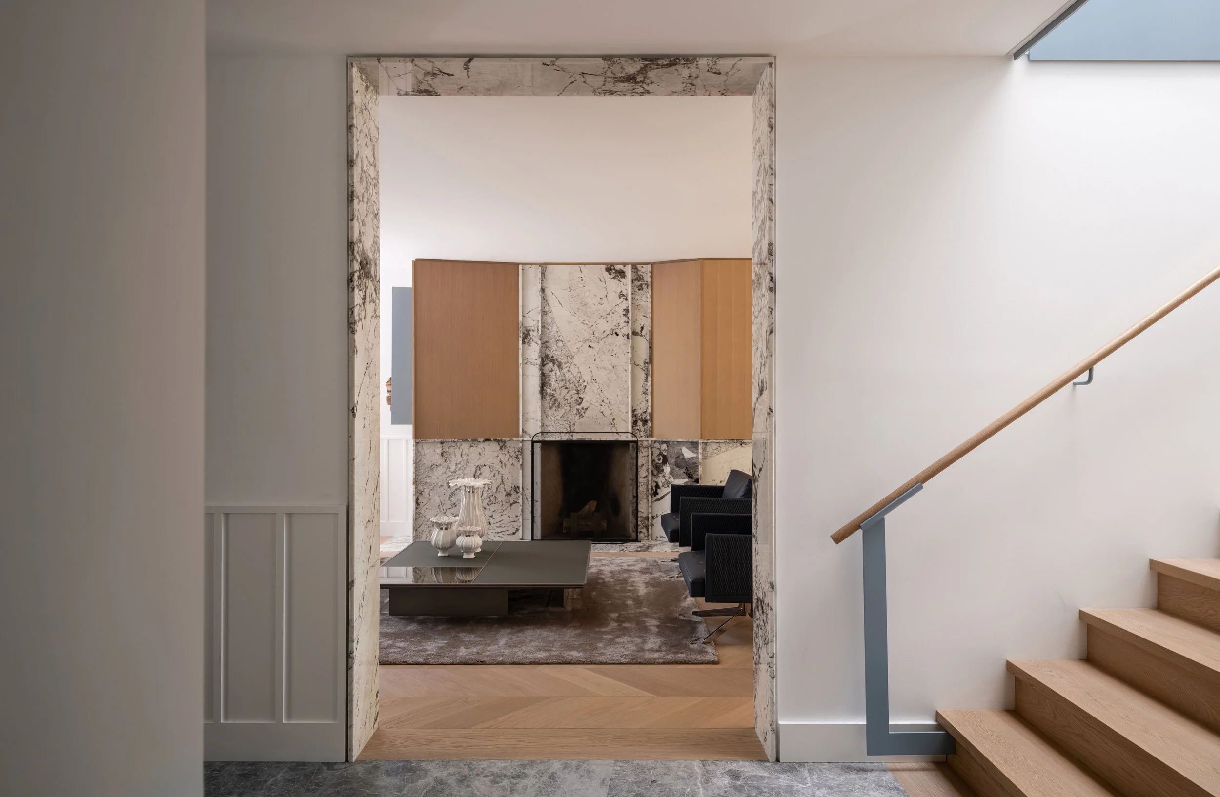 Interior view of a modern living room with a stone fireplace, a marble border, a wood millwork, a dark sofa, and a glass-top coffee table with decorative vases, seen through an archway next to a staircase with wooden steps and a railing.