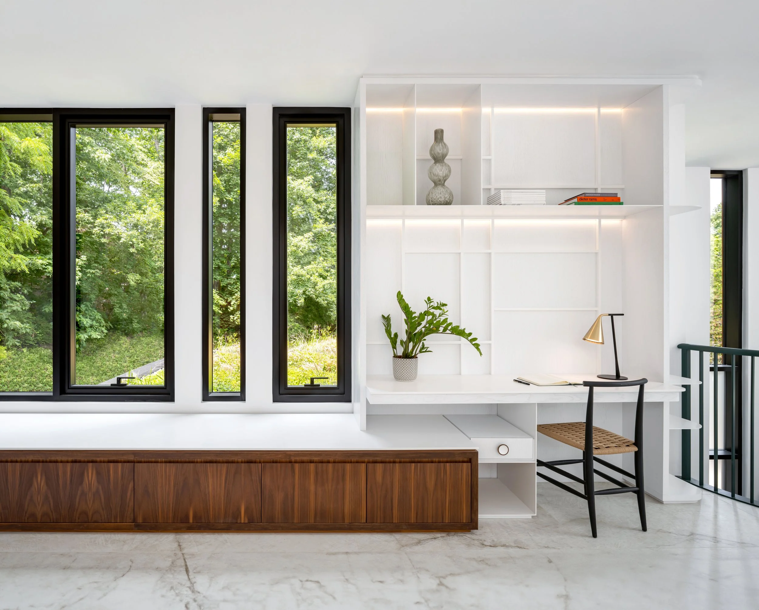 Modern interior with three tall windows overlooking lush greenery, white built-in shelves with decorative items and a potted plant, a small desk with a chair and a desk lamp, and a wooden storage unit at the bottom.