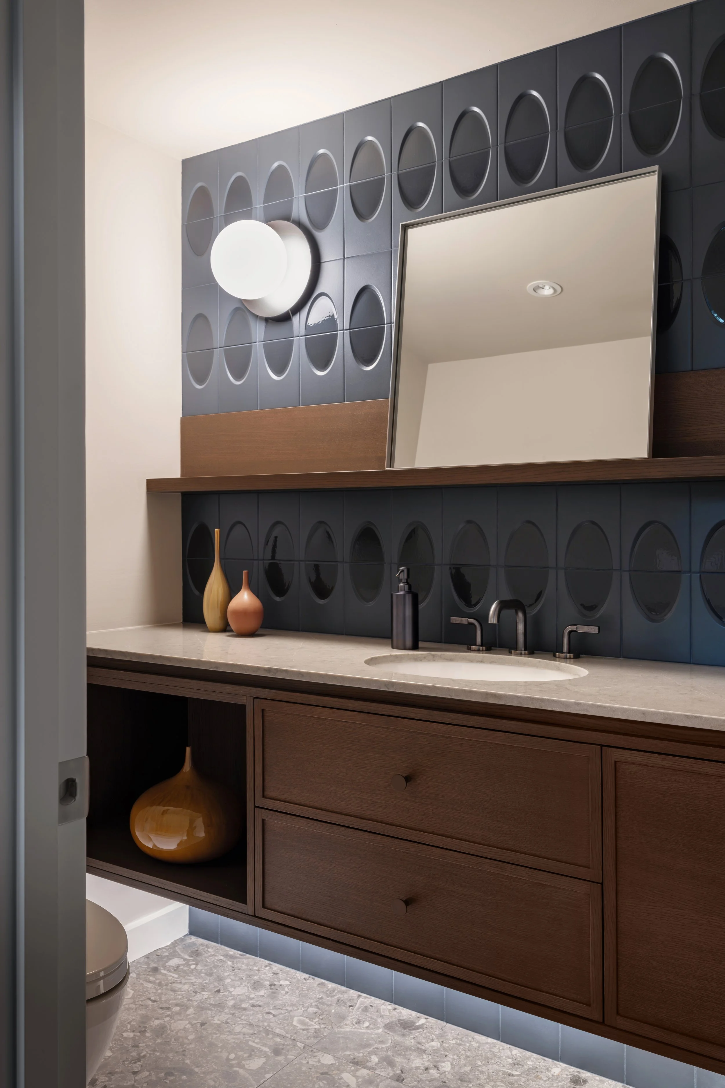 Modern bathroom with blue tile wall, wooden vanity with drawers, a bathroom mirror, and decorative vases on the counter.