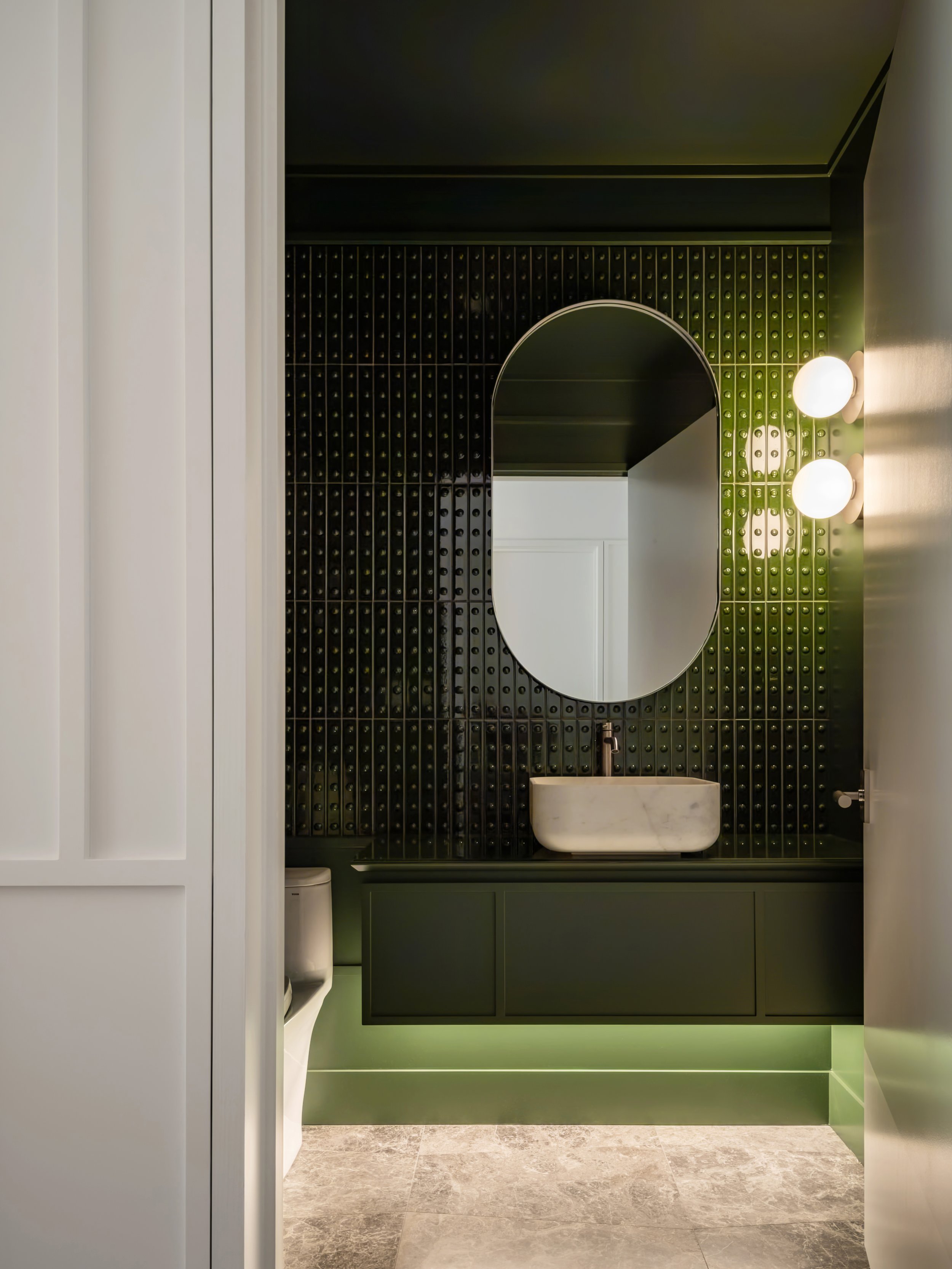A modern bathroom with a large oval mirror on a green tiled wall, green cabinetry, a white marble sink, and wall-mounted globe lights.