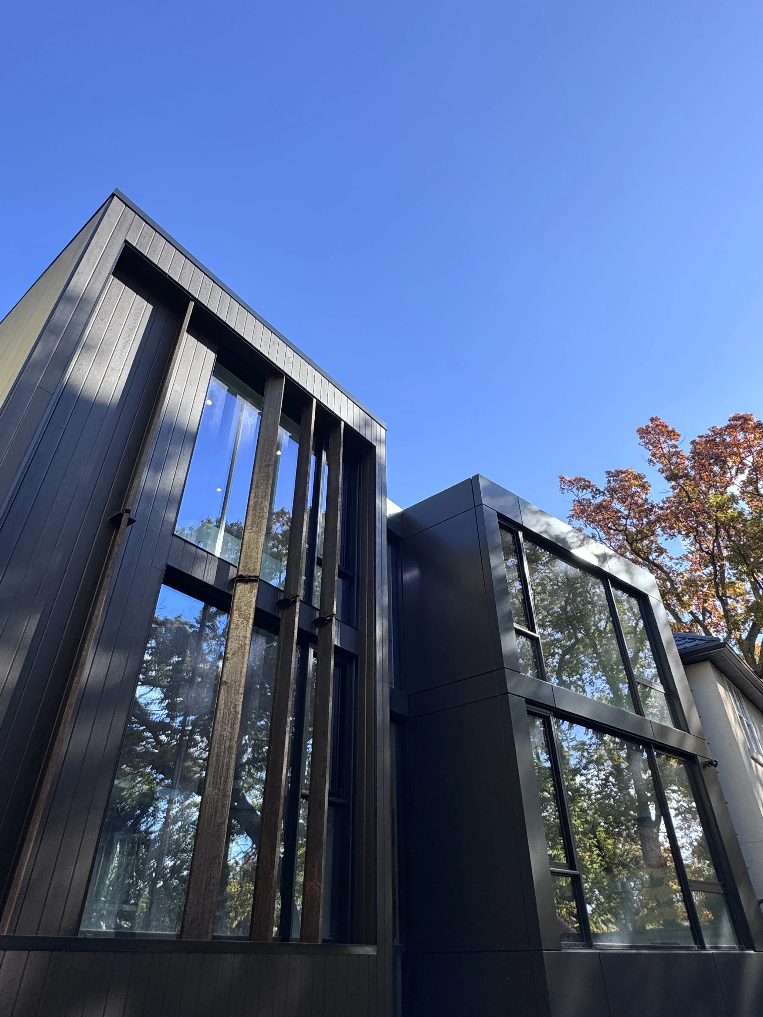 Modern black building with large glass windows, set against a clear blue sky and trees with autumn leaves.