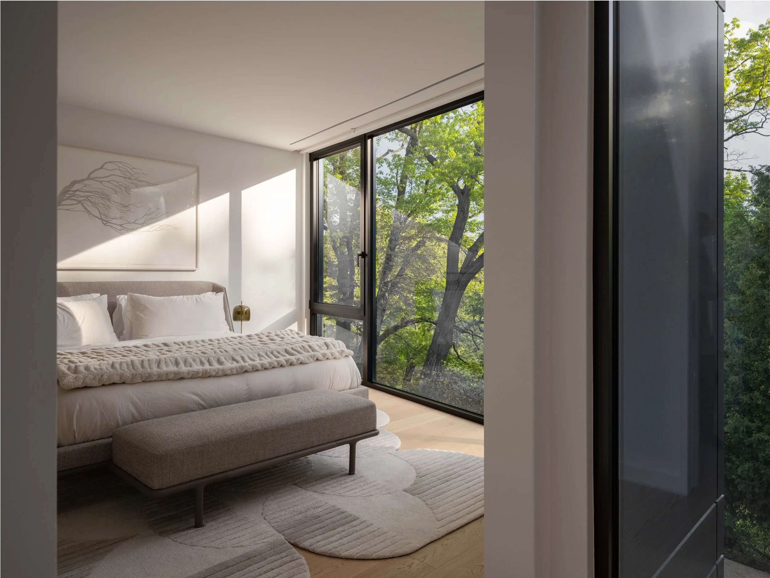 Modern bedroom with large floor-to-ceiling windows showing green trees outside, white walls, a bed with a beige upholstered headboard, white bedding, a gold bedside lamp, and a light-colored rug.