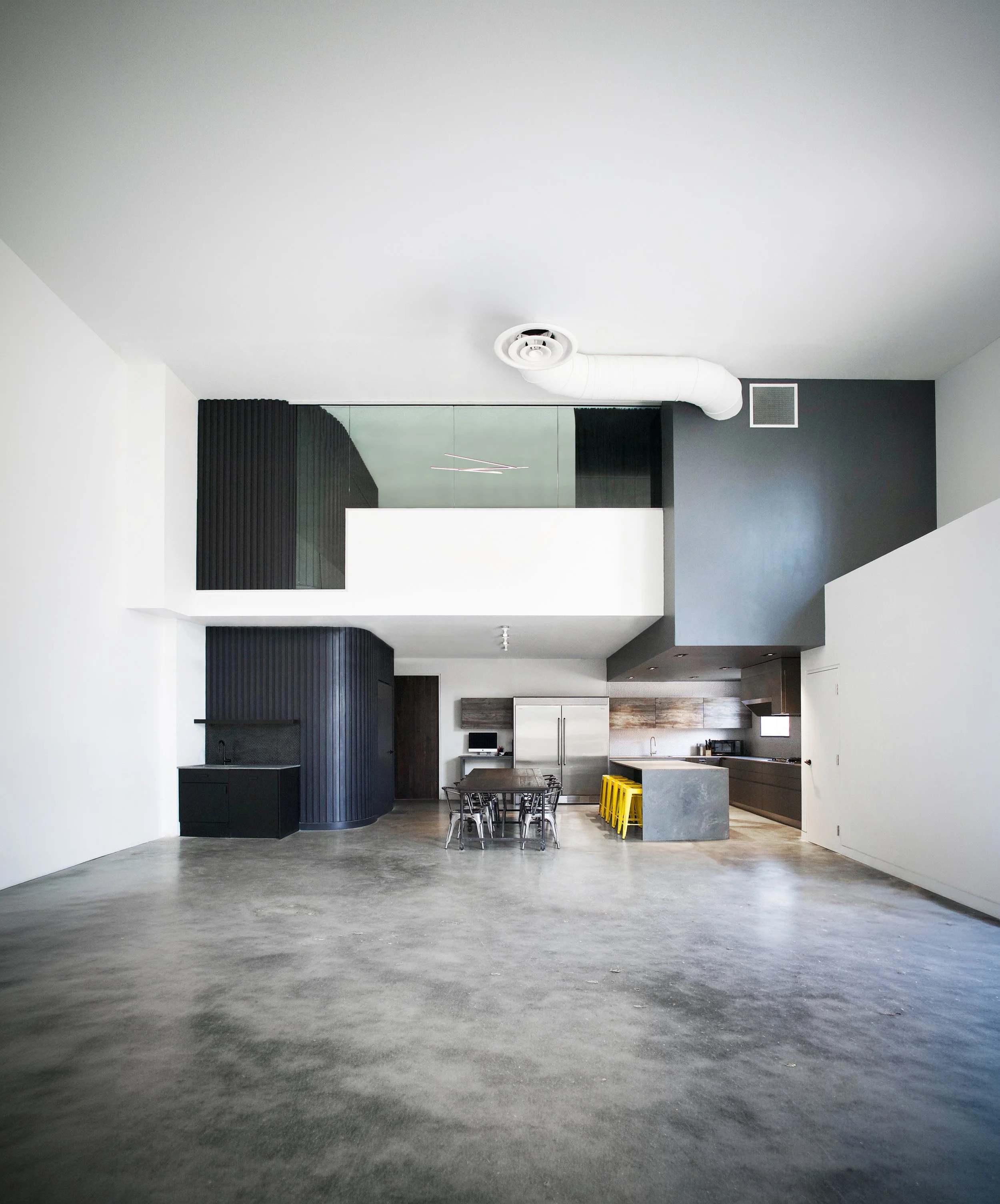 Open-concept modern kitchen and dining area with concrete flooring, white walls, black cabinetry, and a gray island with yellow stools.