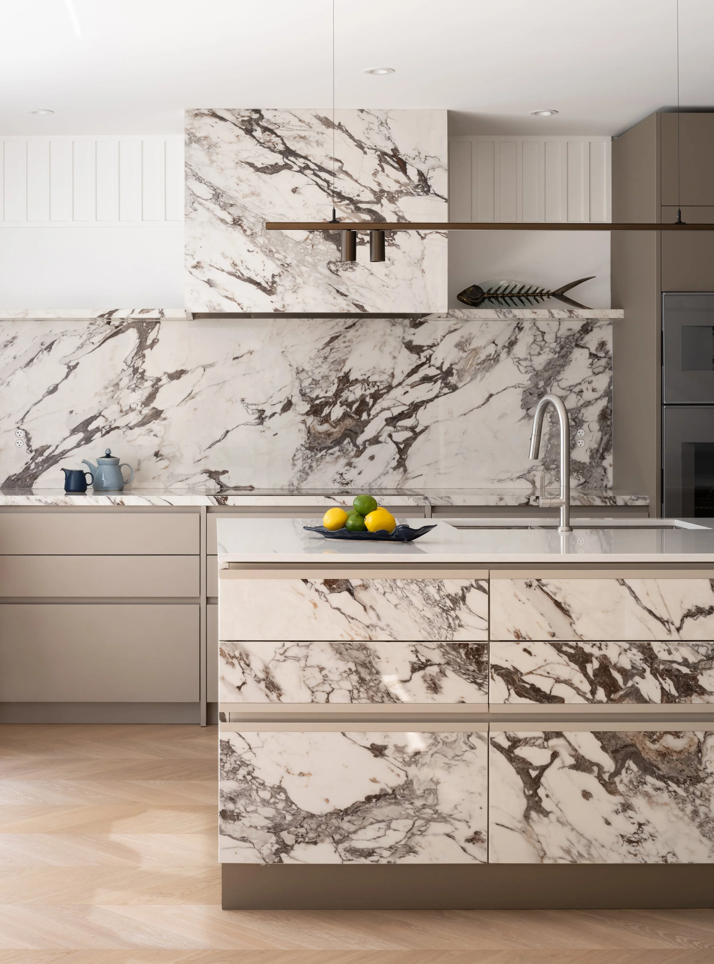 Modern kitchen with marble backsplash and countertops, beige cabinets, a sink with a silver faucet, a fishbone sculpture on a shelf, and a blue tray with lemons and limes.