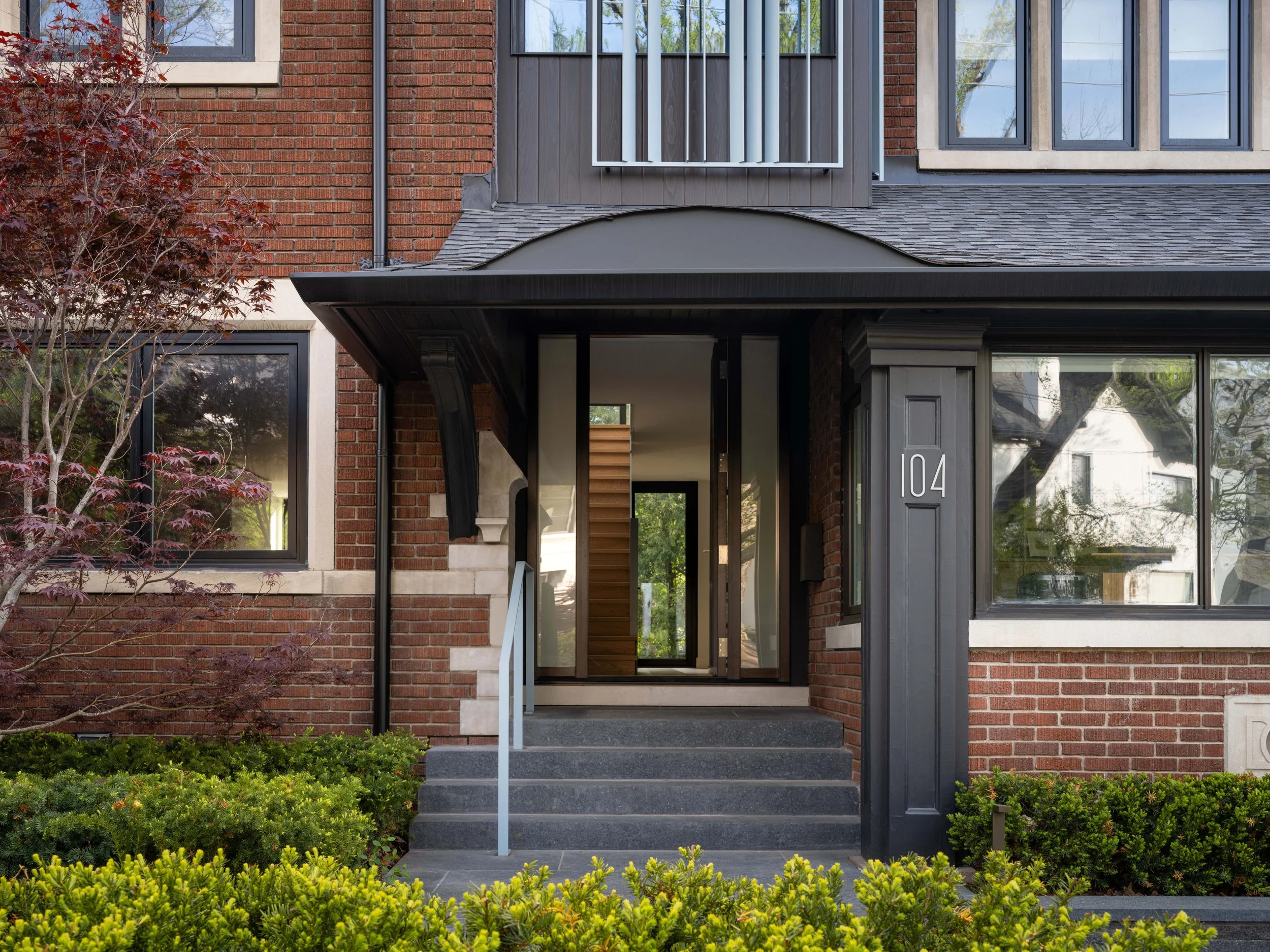 Front view of a modern brick house with large windows, a small covered porch, black trim, gray granite steps, and surrounding green shrubs and trees.