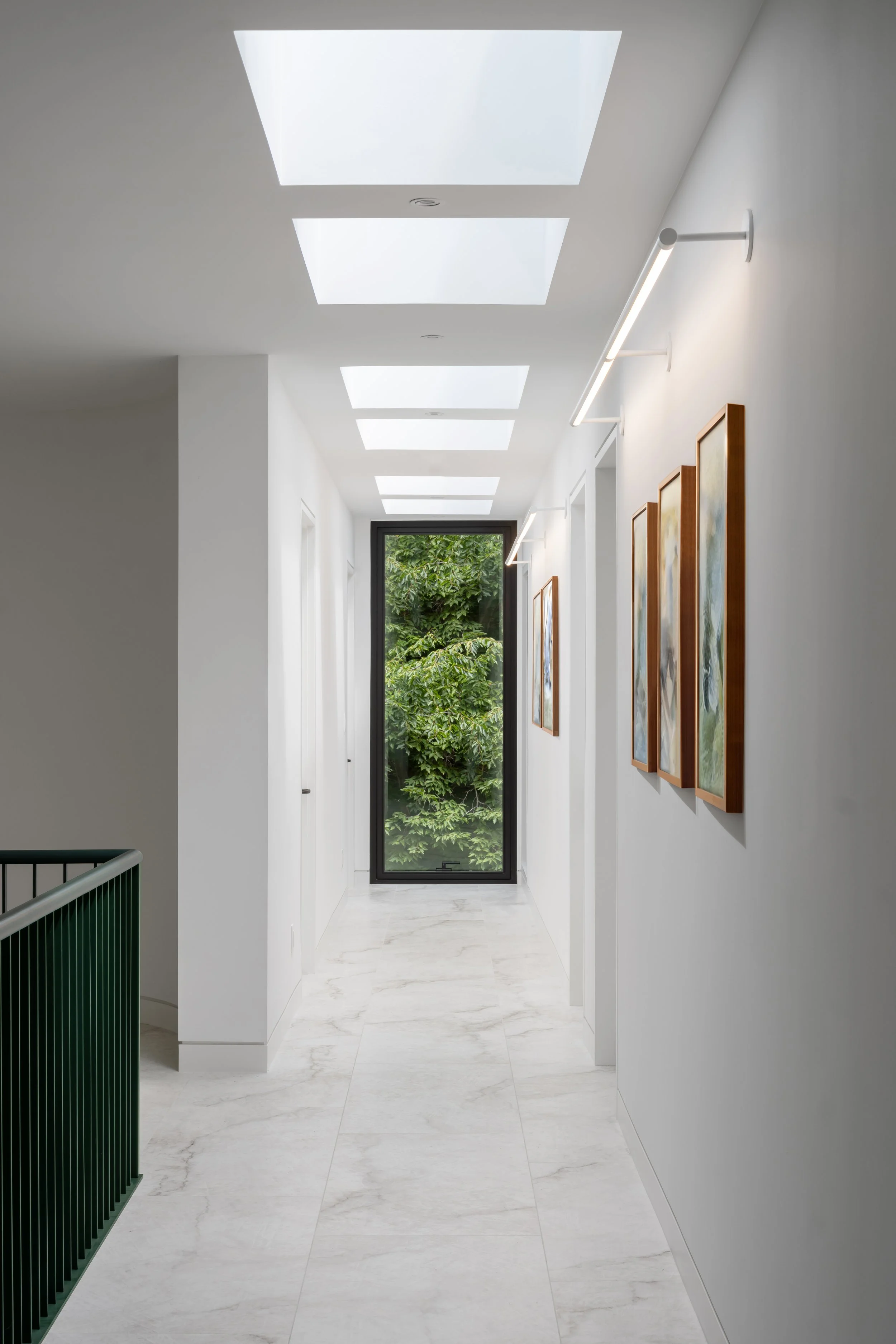 A clean, modern hallway with white walls, marble flooring, three skylights, framed artwork on the right wall, a large window at the end showing green trees, and a green railing on the left side.