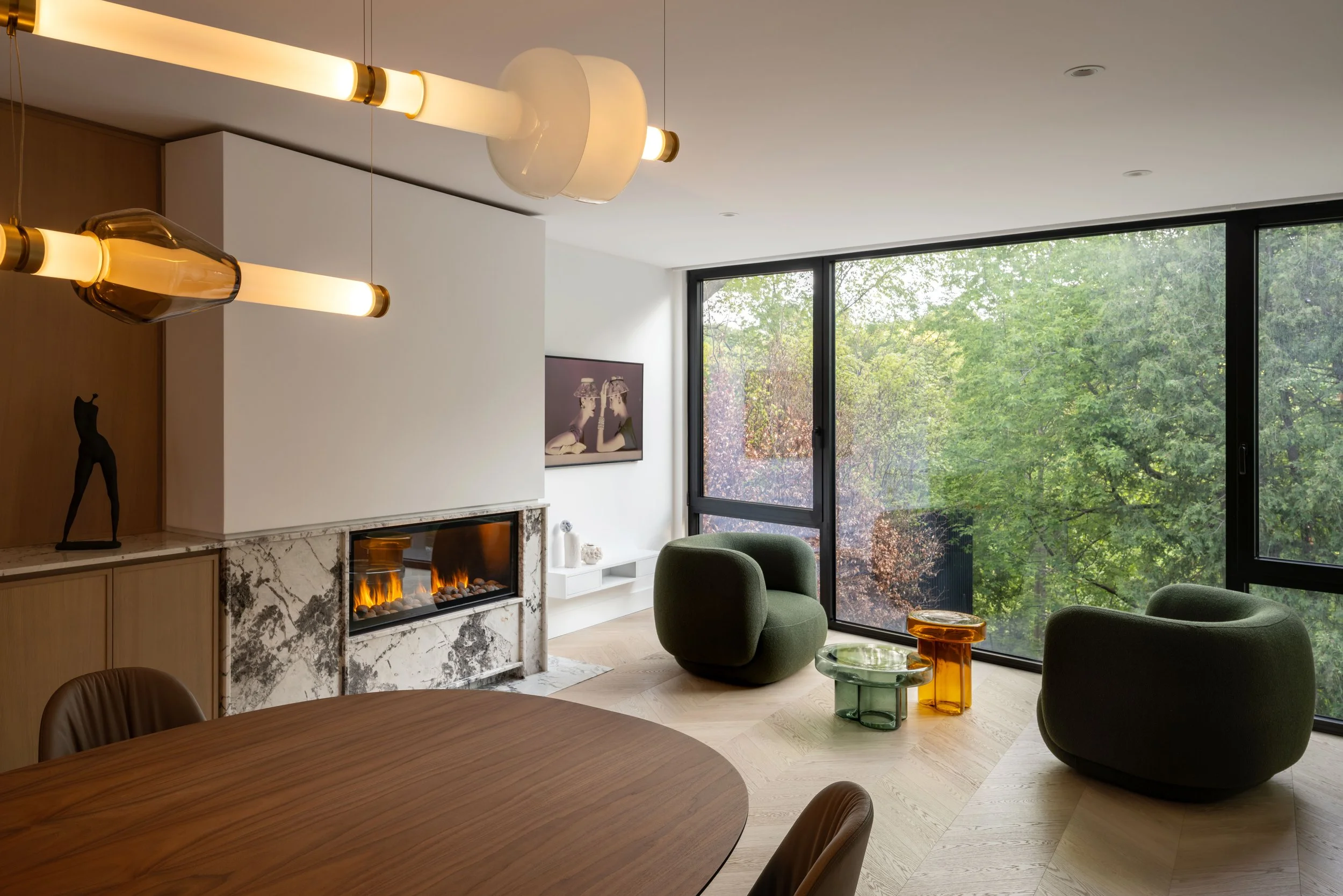 Modern living room with large windows overlooking green trees, a fireplace, green chairs, wood dining table, and abstract glass chandelier.