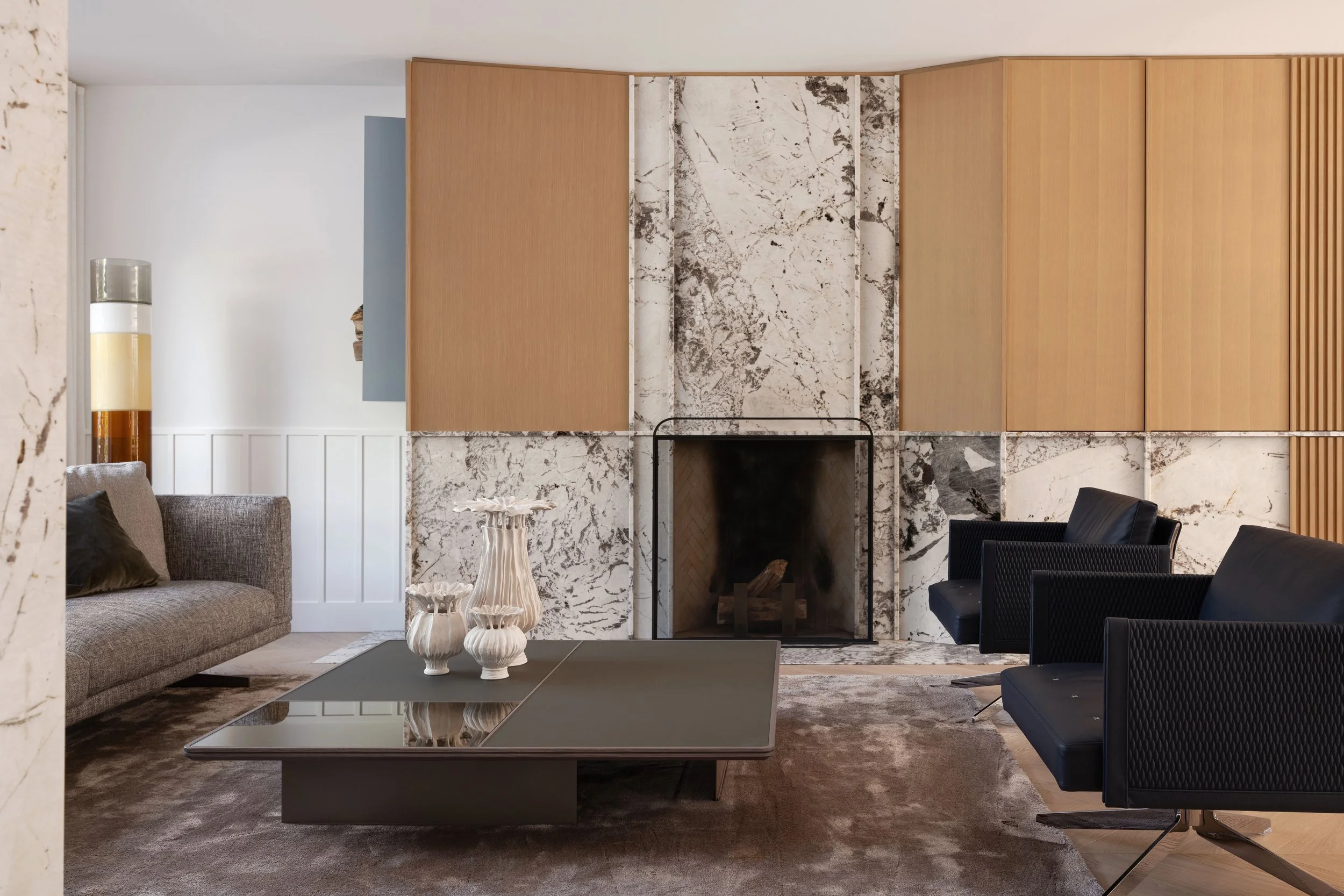 Living room with a brown sofa, blue chairs, marble fireplace, marble and wood millwork panels, patterned vases, and a coffee table.