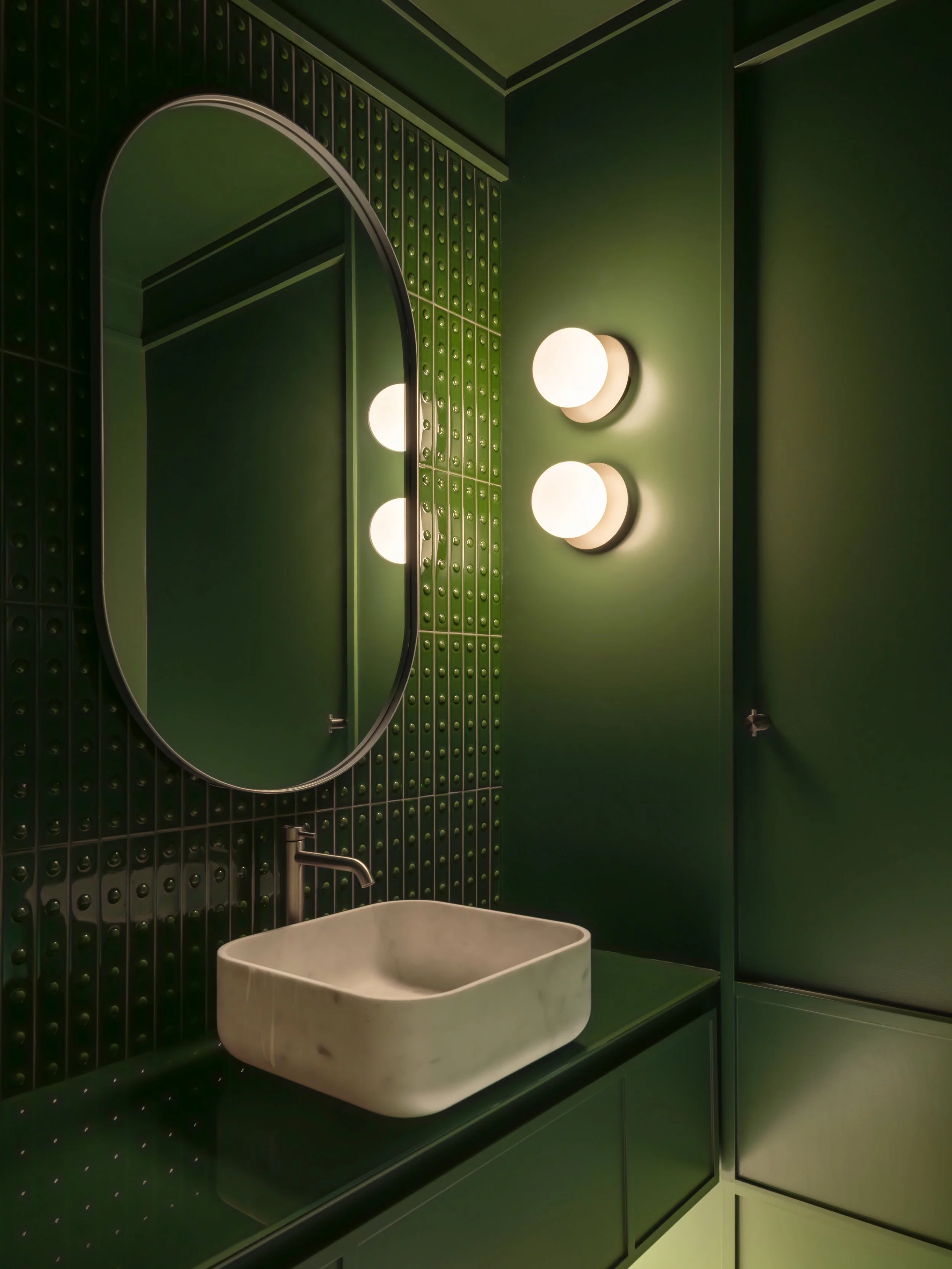 A modern bathroom with dark green tiled walls, a large oval mirror, two spherical wall lights, a white marble sink, and green custom vanity.