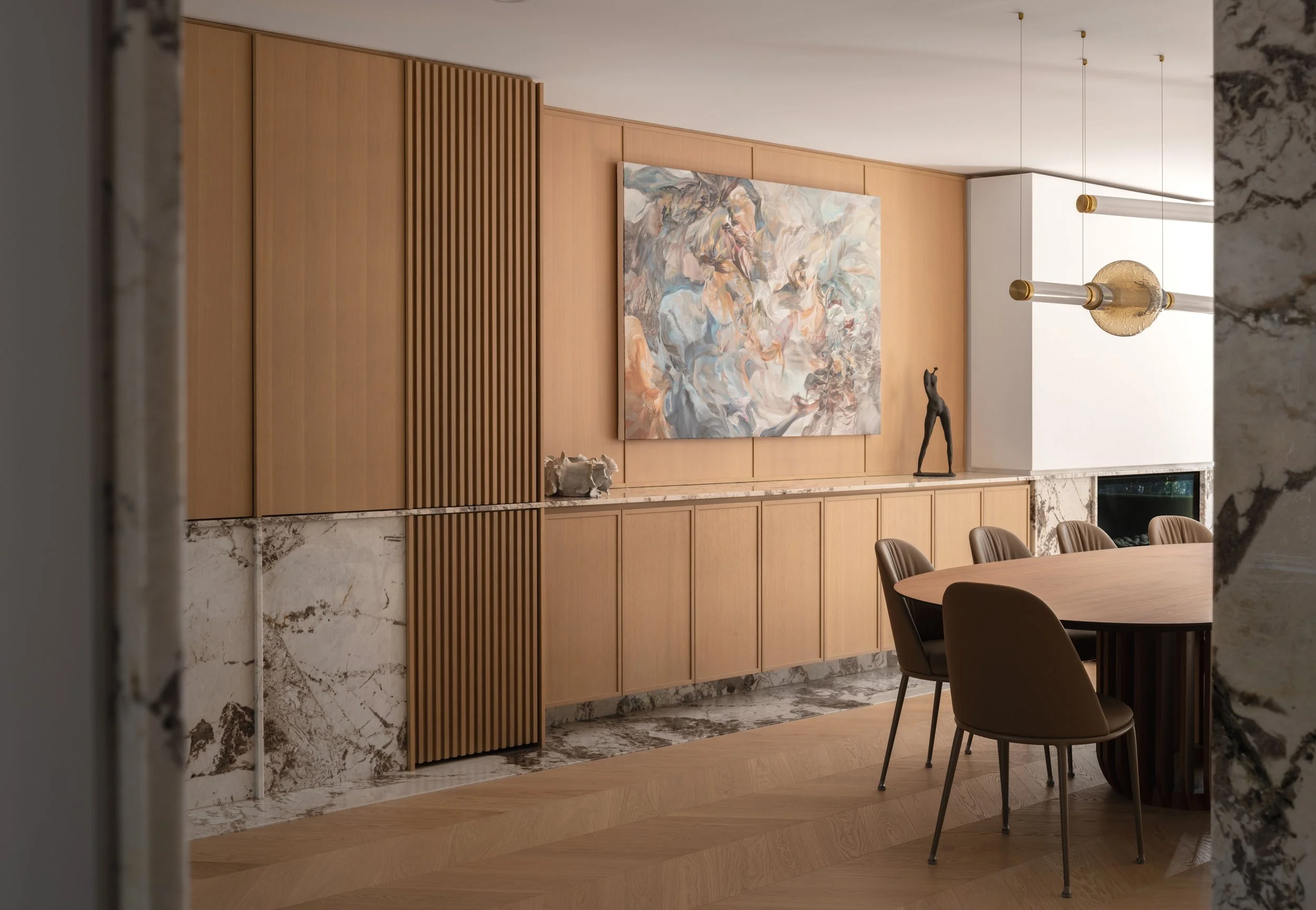 A modern dining room with a large wooden wall panel, a marble fireplace, and a large abstract painting. Dining table with brown chairs, and hanging light fixture.