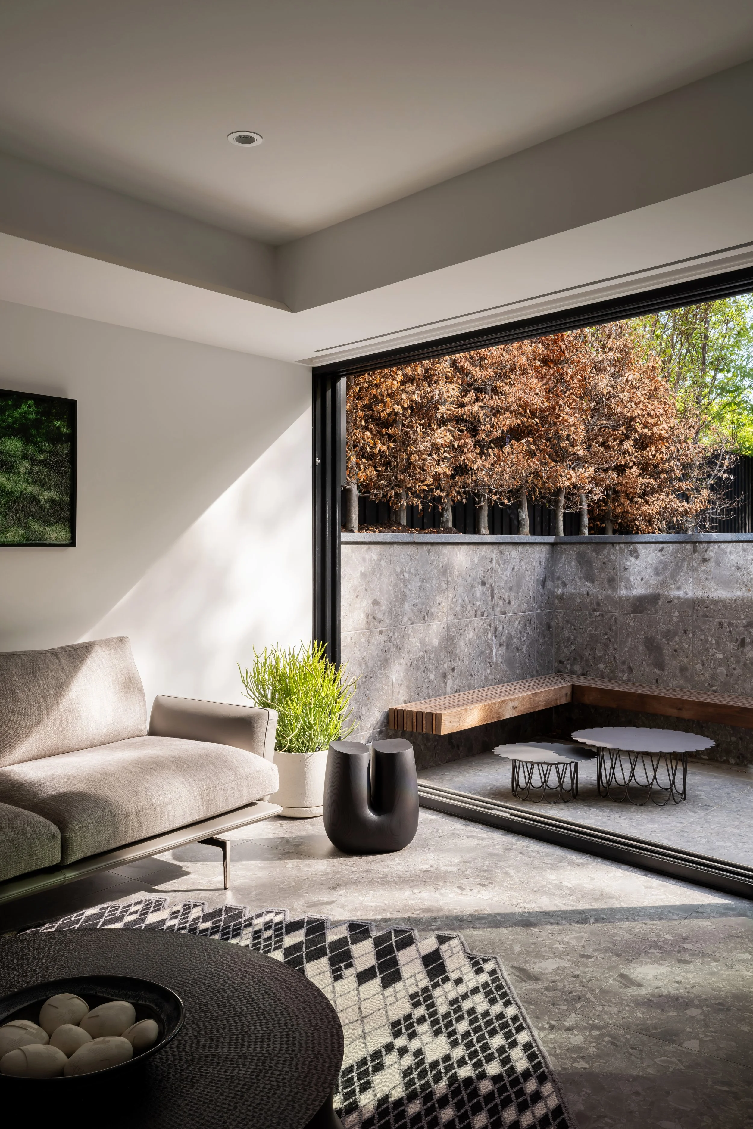 Interior living space with a beige sofa, a potted plant, black vases, geometric-patterned rug, and a sliding glass door leading to an outdoor area with trees.