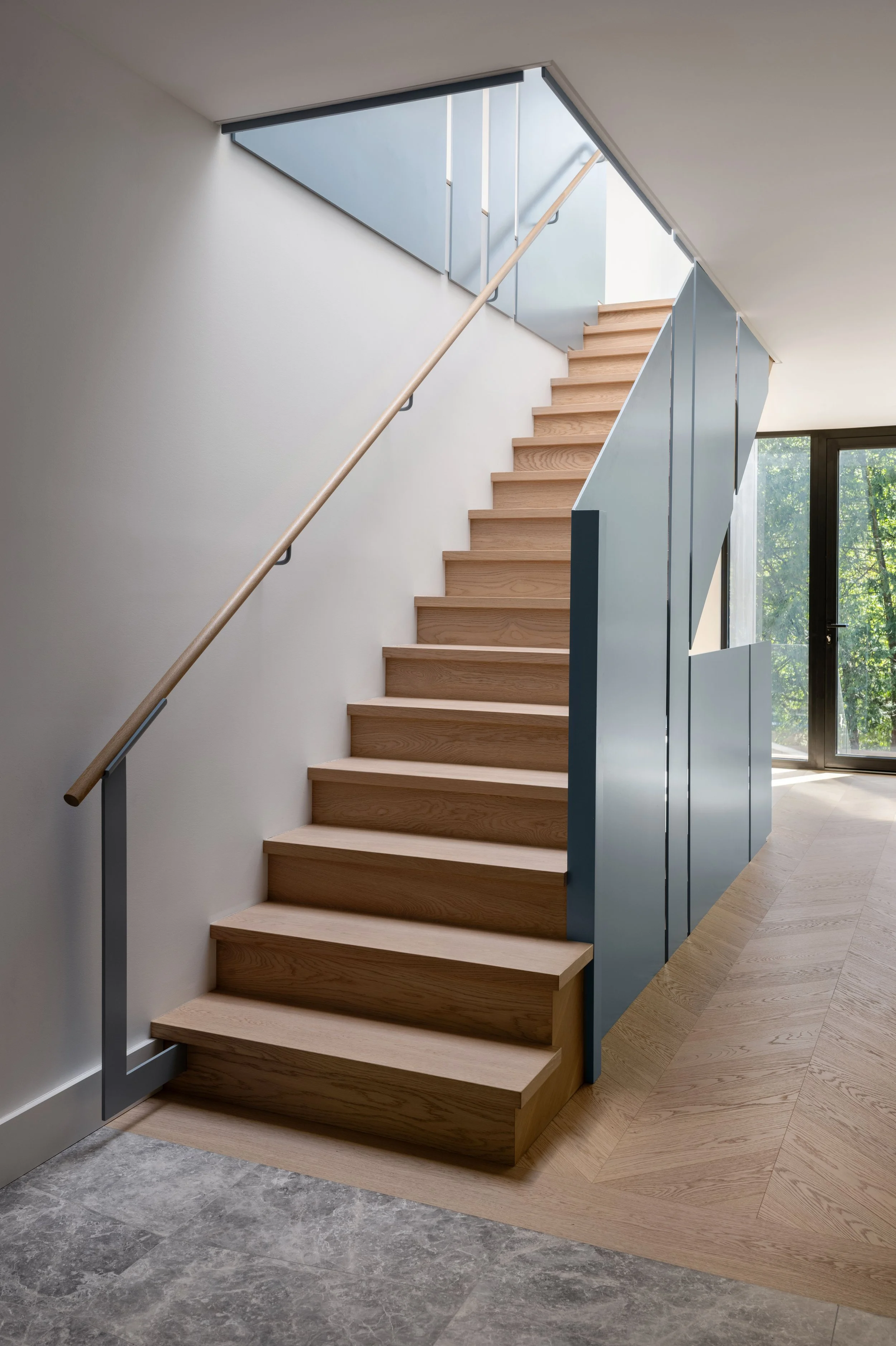Modern staircase with wooden steps, wood handrail, and blue steel panels in a contemporary interior with large window and mixed flooring.