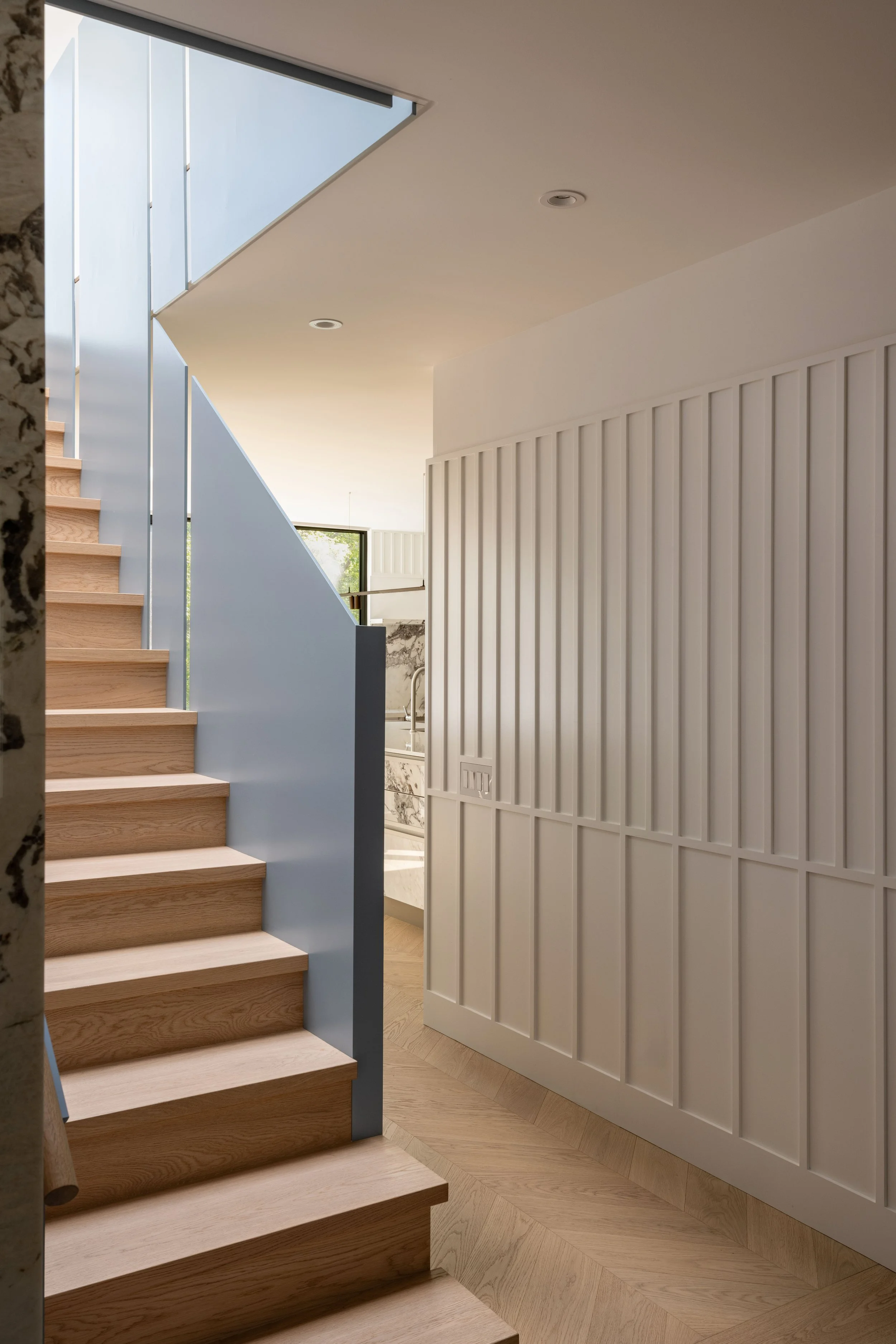 Interior view of a modern staircase with light wooden steps, a blue staircase guard, and a beige wall with vertical paneling. There is a window in the background allowing natural light into the space.