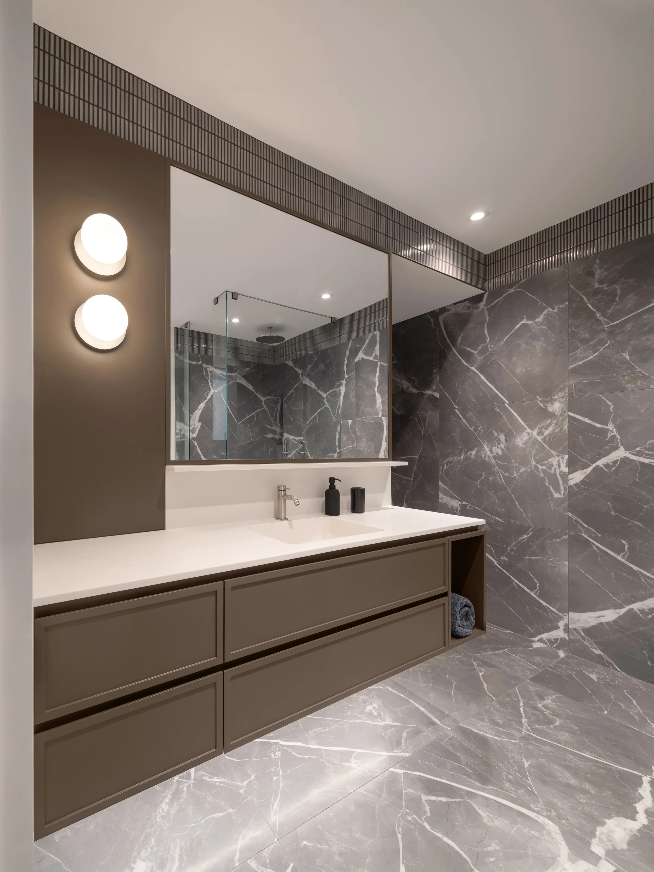 A modern bathroom with a brown vanity, a large rectangular mirror, black soap dispensers, gray porcelain wall tiles, and matching porcelain floor tiles. There are minimalist wall-mounted light fixtures and a glass shower enclosure in the reflection.