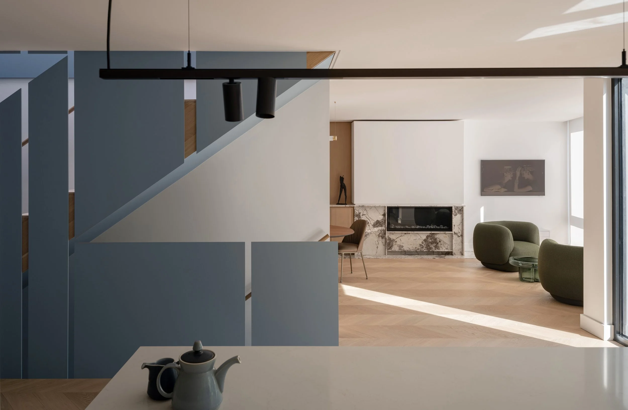 Modern living room with a light wood floor, green armchair, and a marble fireplace. To the left, a blue steel staircase  leads upstairs. Natural light streams through large windows.