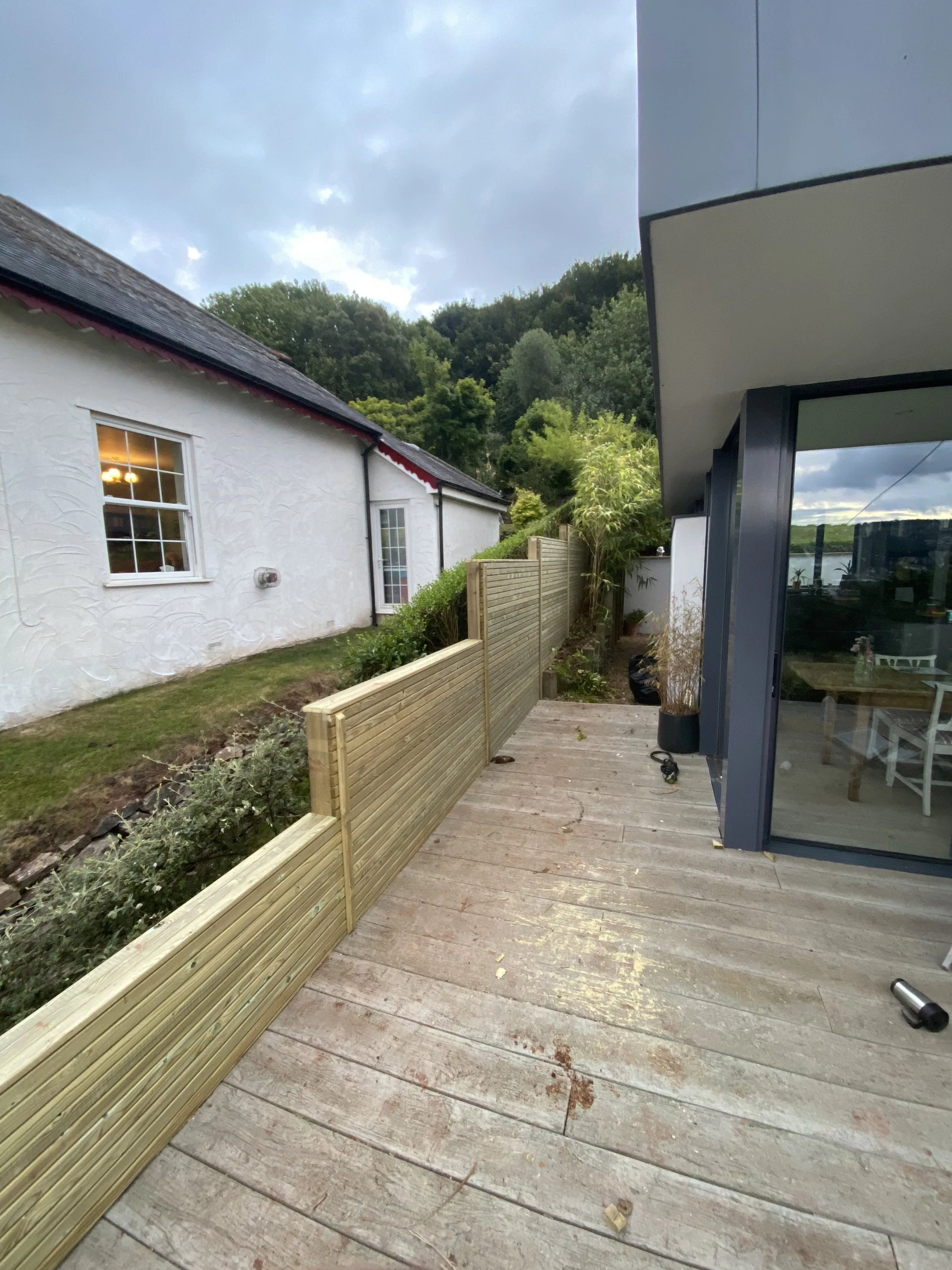 View of a backyard patio with wooden decking, a new wooden privacy fence, outdoor plants, and a white house with windows, under a cloudy sky.