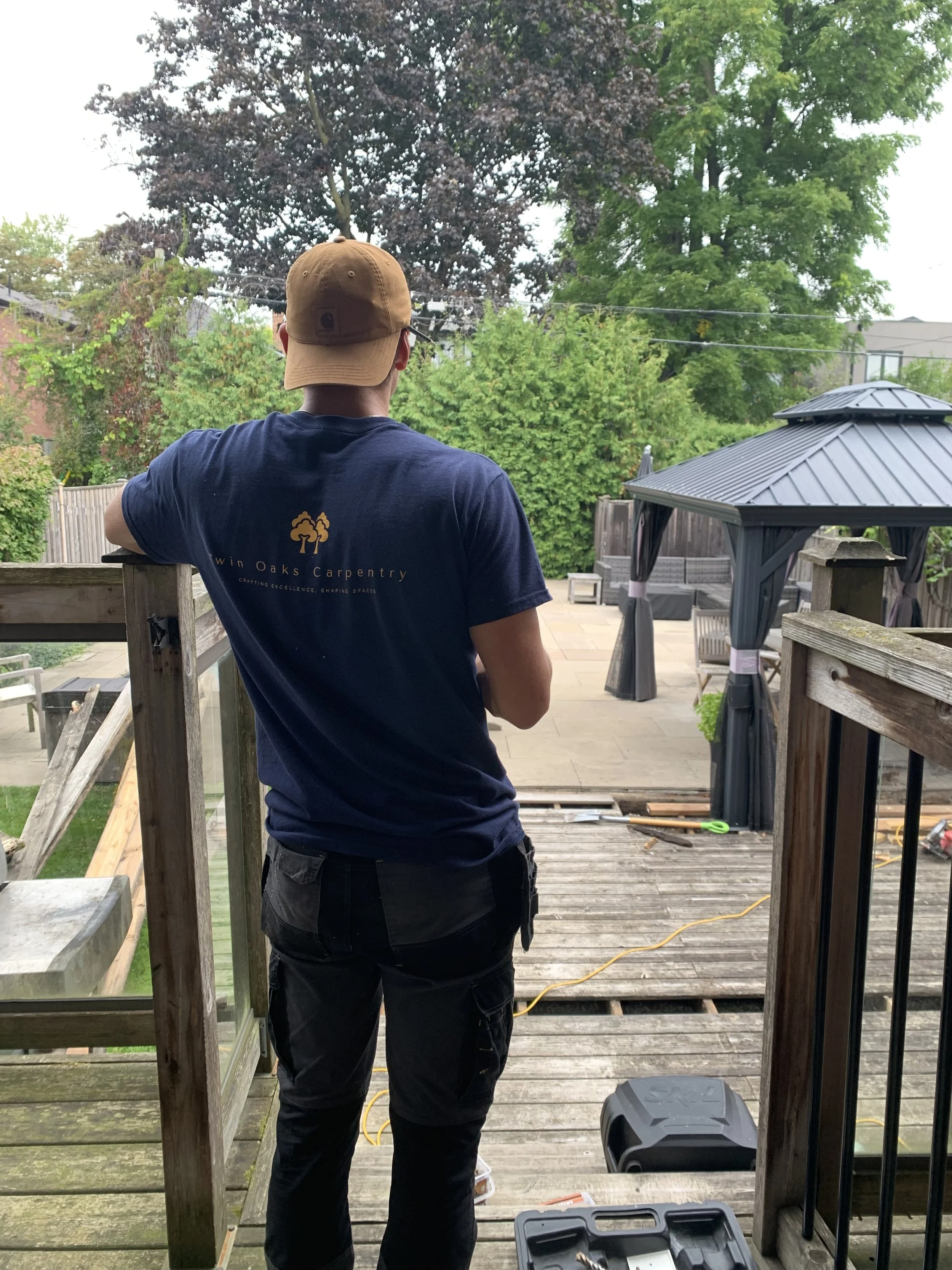 Person standing on the wooden deck, wearing a brown cap and a navy blue t-shirt with a tree logo and text, overlooking a backyard with trees, a gazebo, and outdoor furniture.