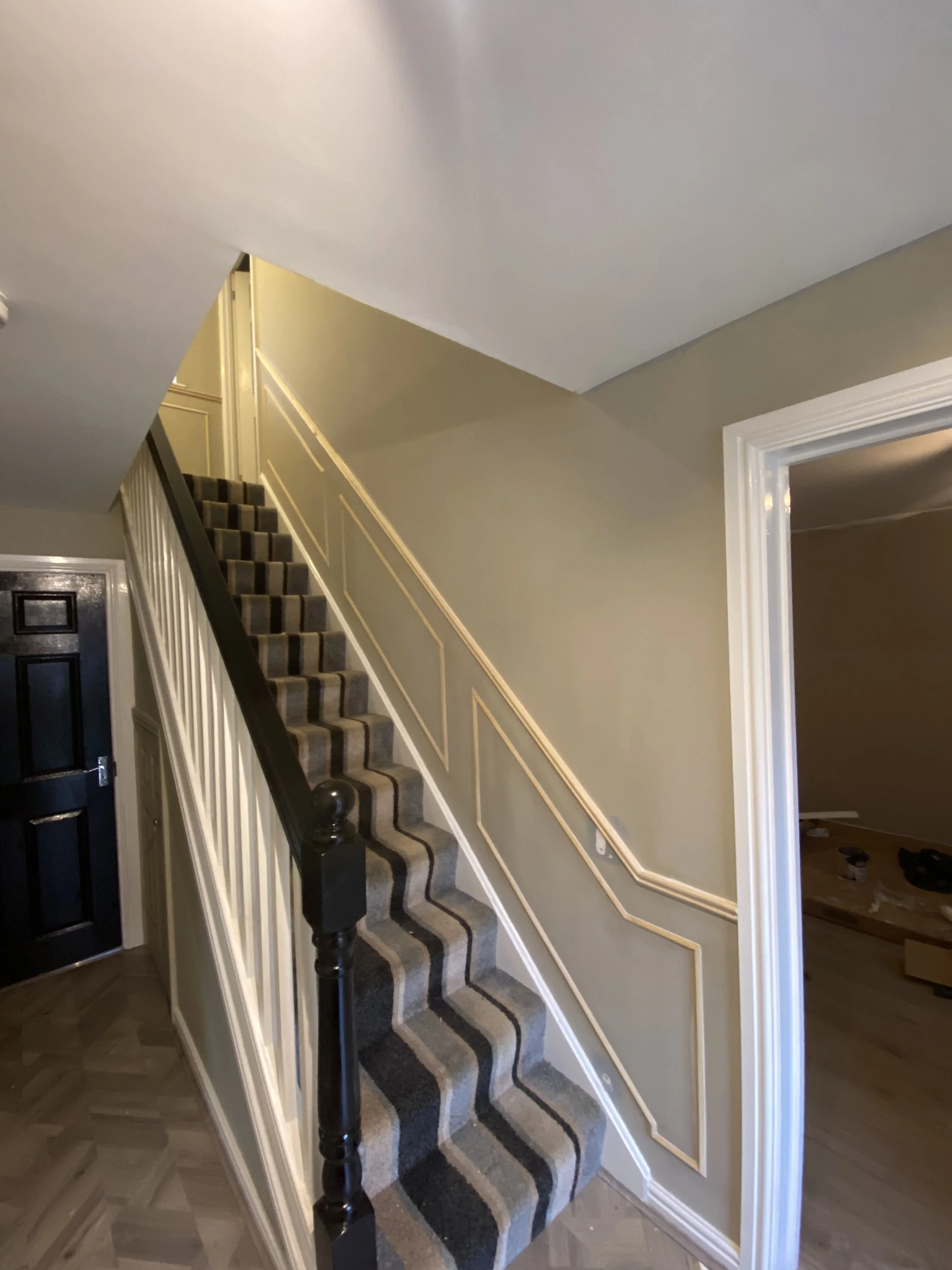 Interior view of a house staircase with a black and gray patterned carpet, white railings, beige walls with decorative molding, and an open doorway leading to another room.