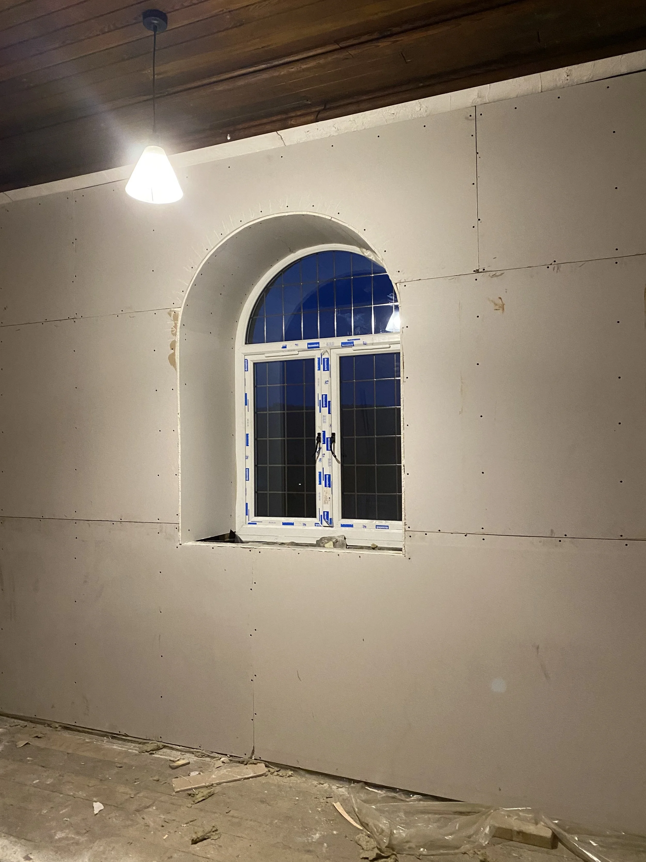 Interior wall under construction with a window and a wooden ceiling, construction debris on the floor, and a ceiling light turned on.