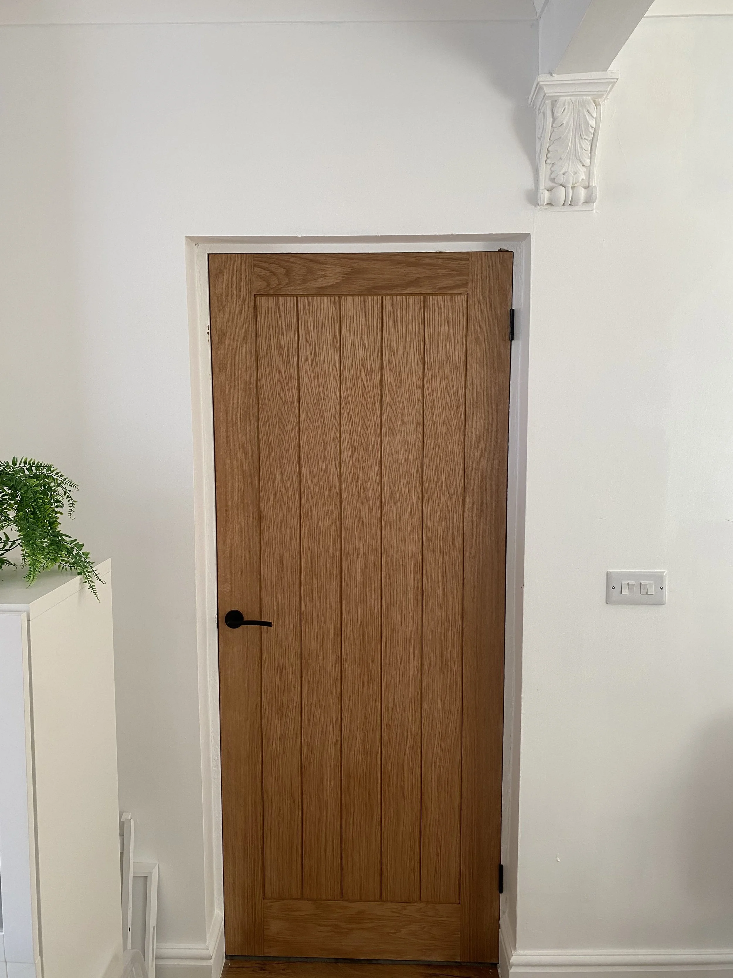 A wooden door with vertical panels and a black handle in a white frame, set in a white wall with ornate crown molding at the top right corner.