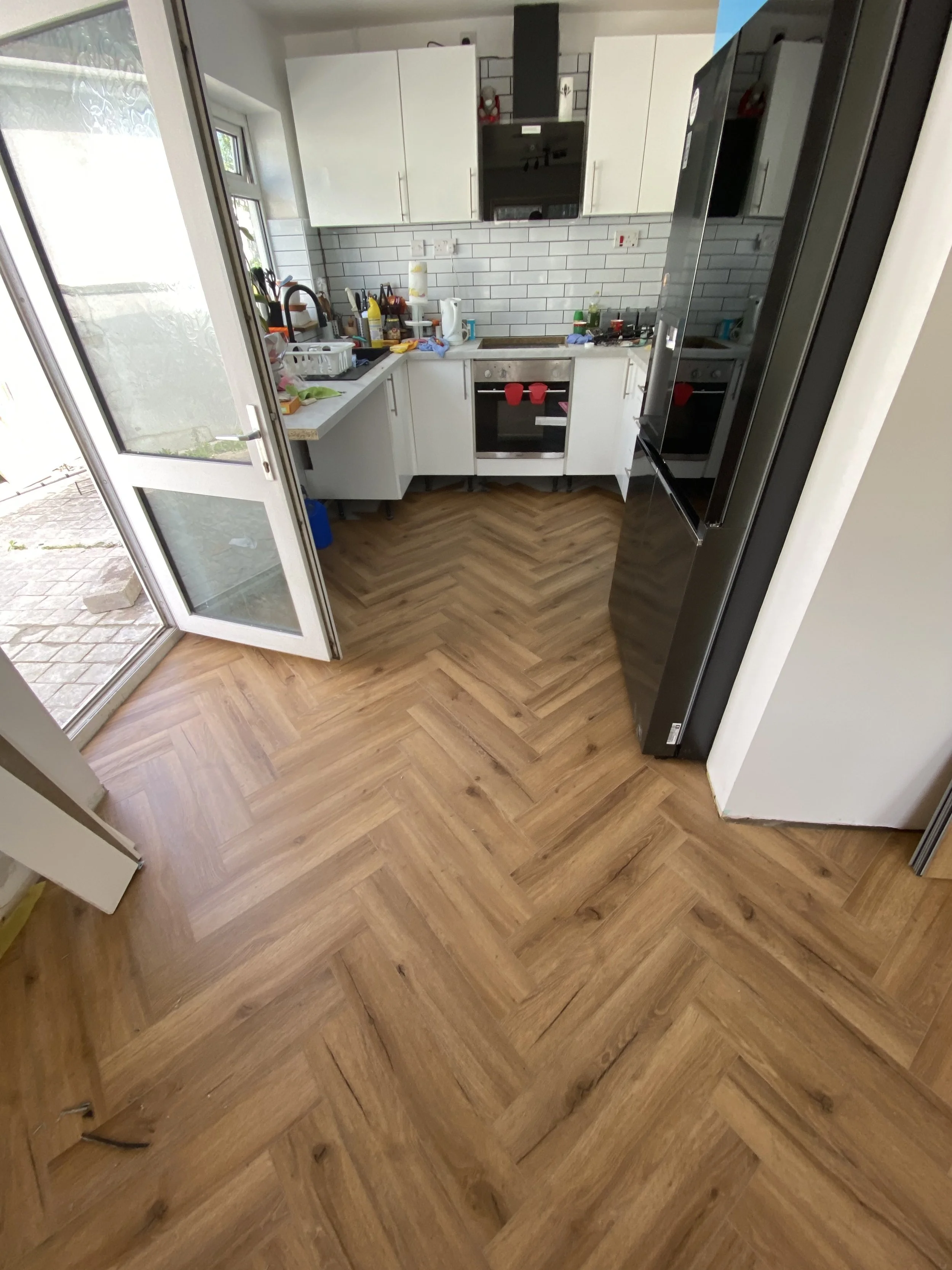 A small kitchen with white cabinets, undermounted sink, and a black oven. There is a refrigerator on the right and a door leading outside on the left. The kitchen has a wood-patterned floor arranged in a herringbone pattern. Countertops are cluttered